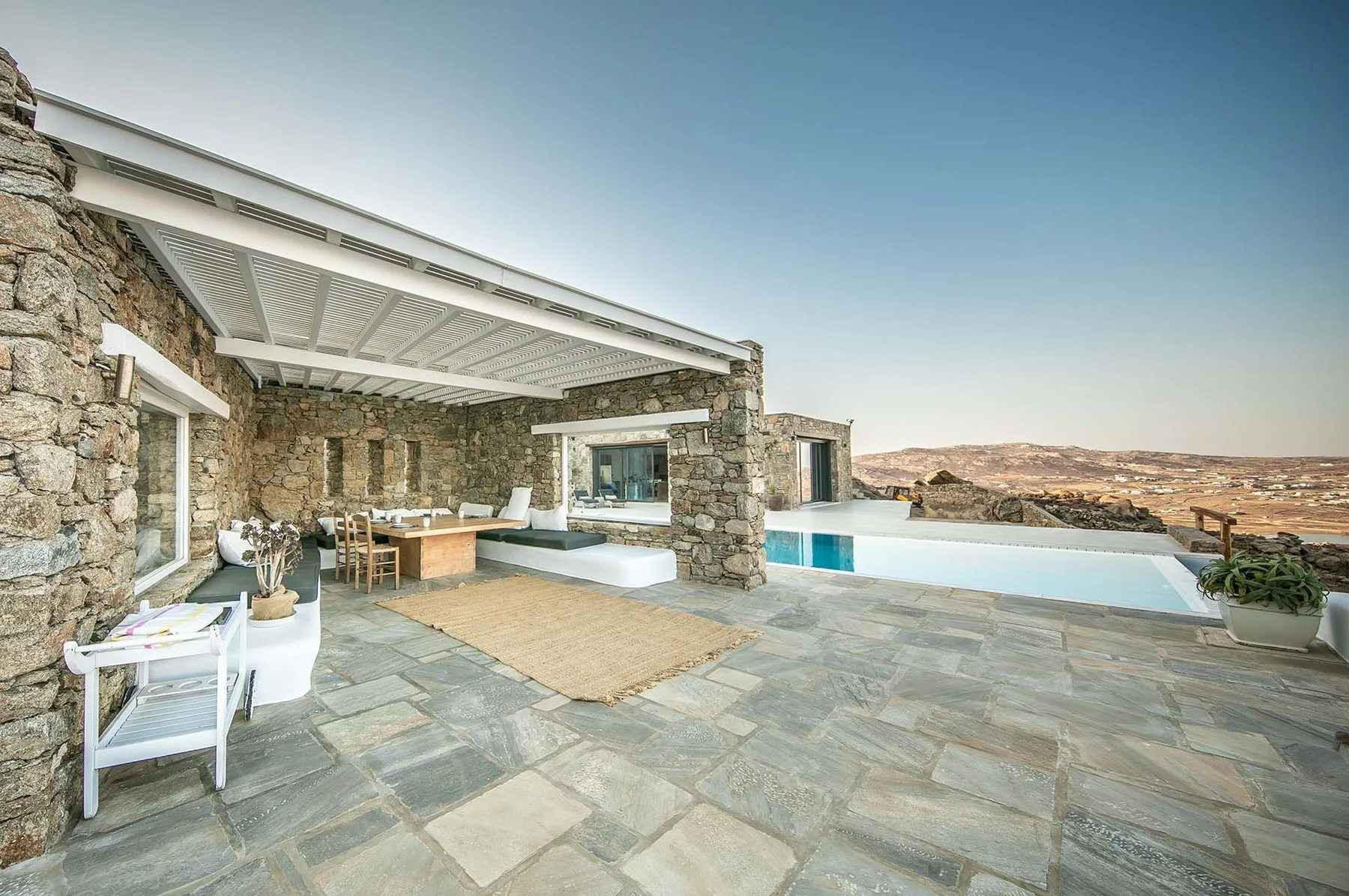 A stone patio with a dining area beside a swimming pool and open landscape.