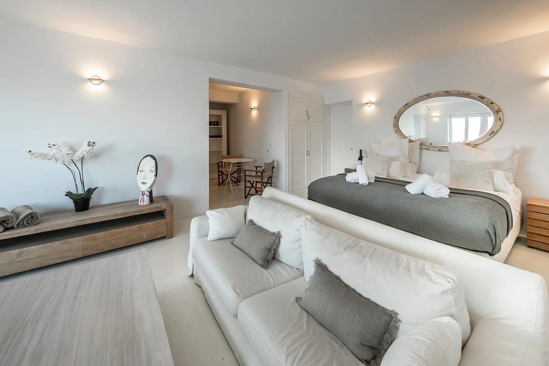 A bright bedroom with a bed, white seating, and an oval mirror.