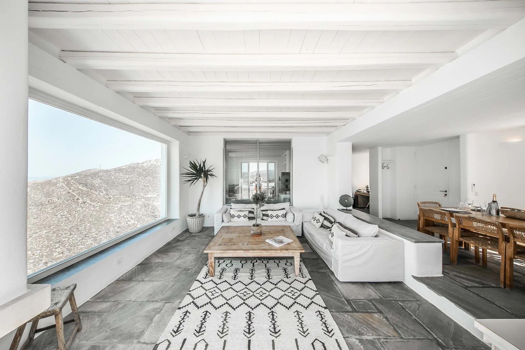 A bright living and dining area has white seating, a low table, and a large window.