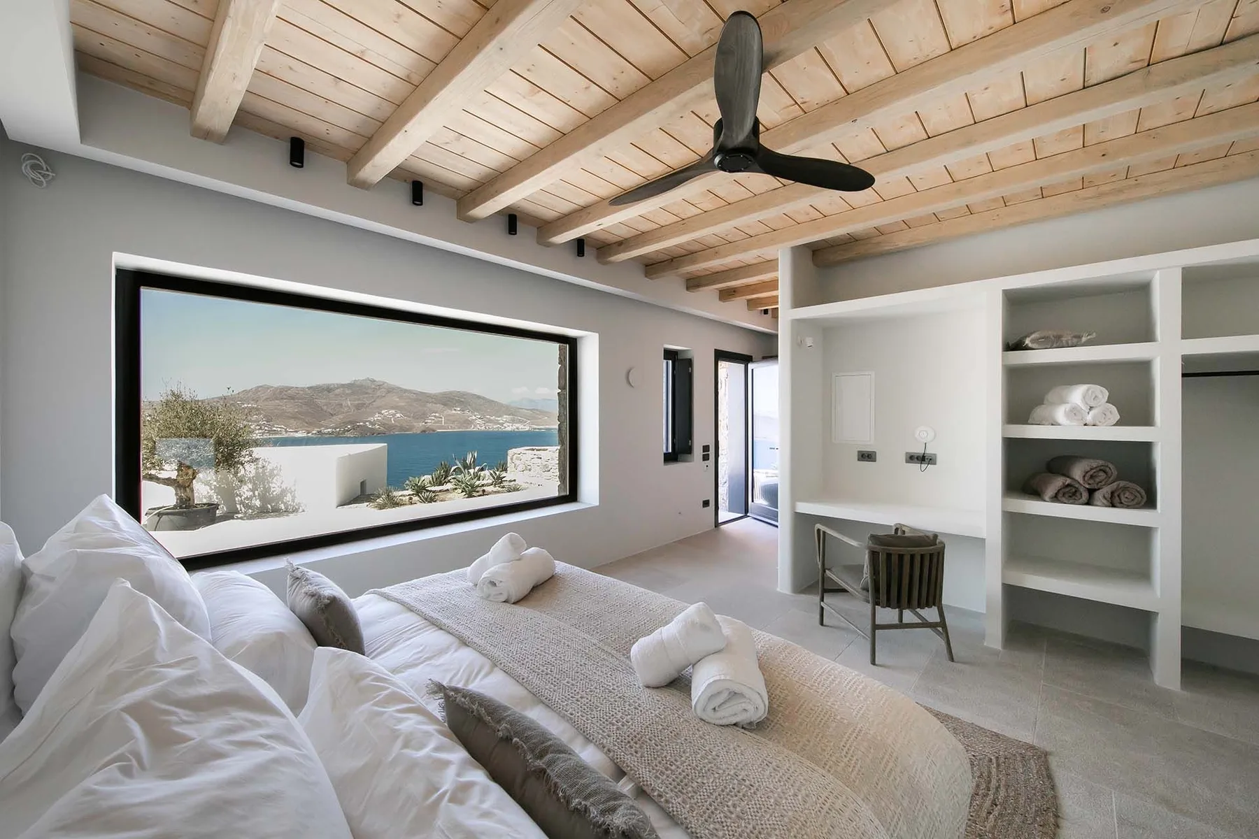 A bright bedroom with white bedding, built-in shelves, and a large window overlooking the water.