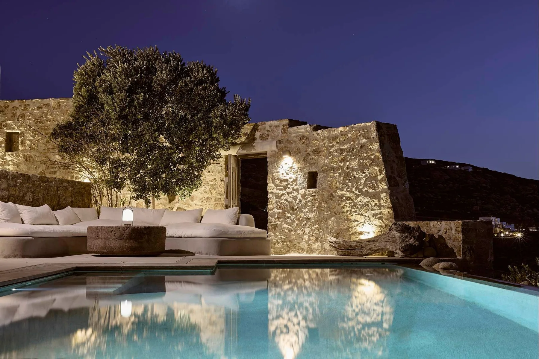 Una piscina exterior iluminada está junto a un edificio de piedra y asientos acolchados de noche.