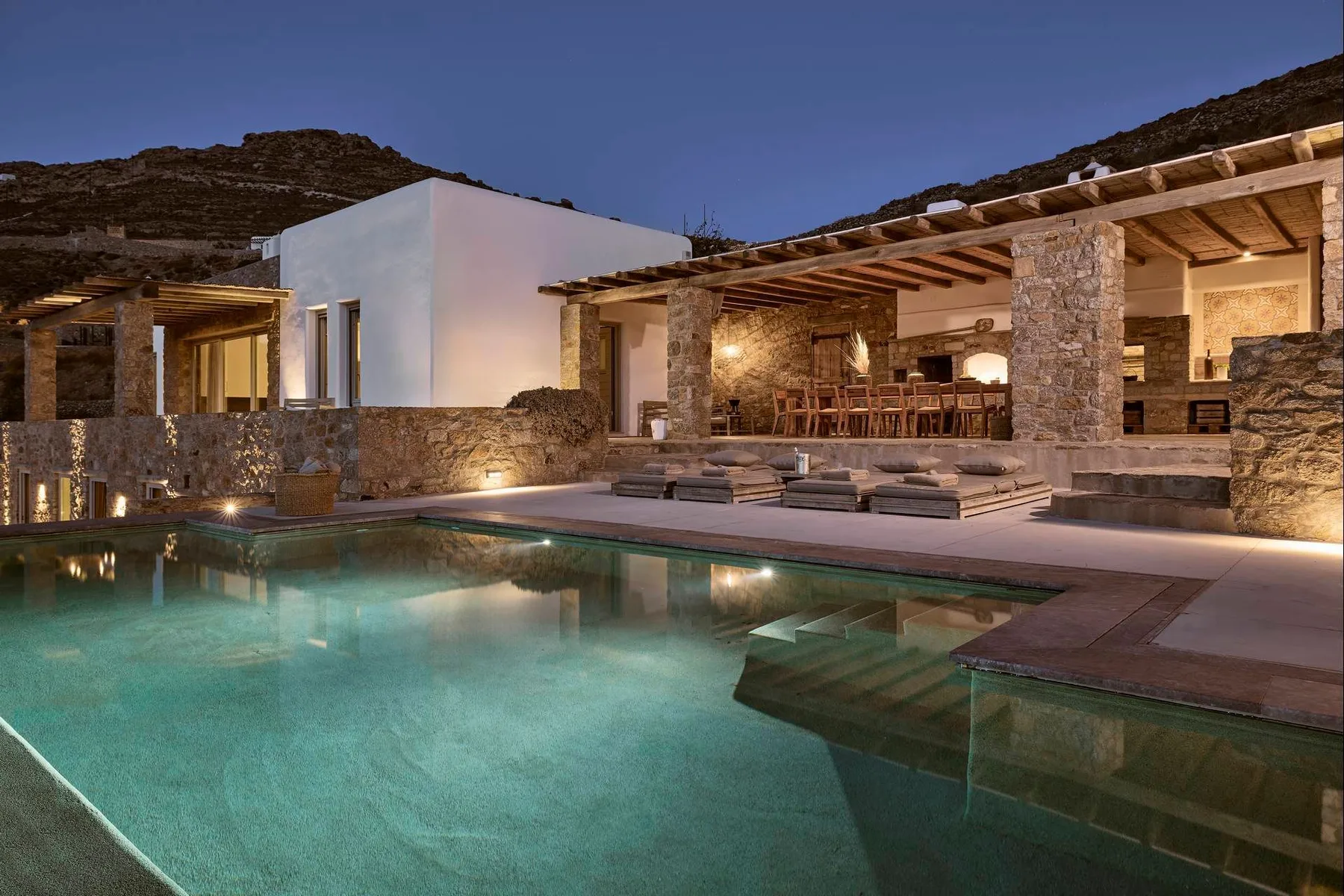 Una piscina junto a una zona exterior de comedor de piedra y madera al anochecer.