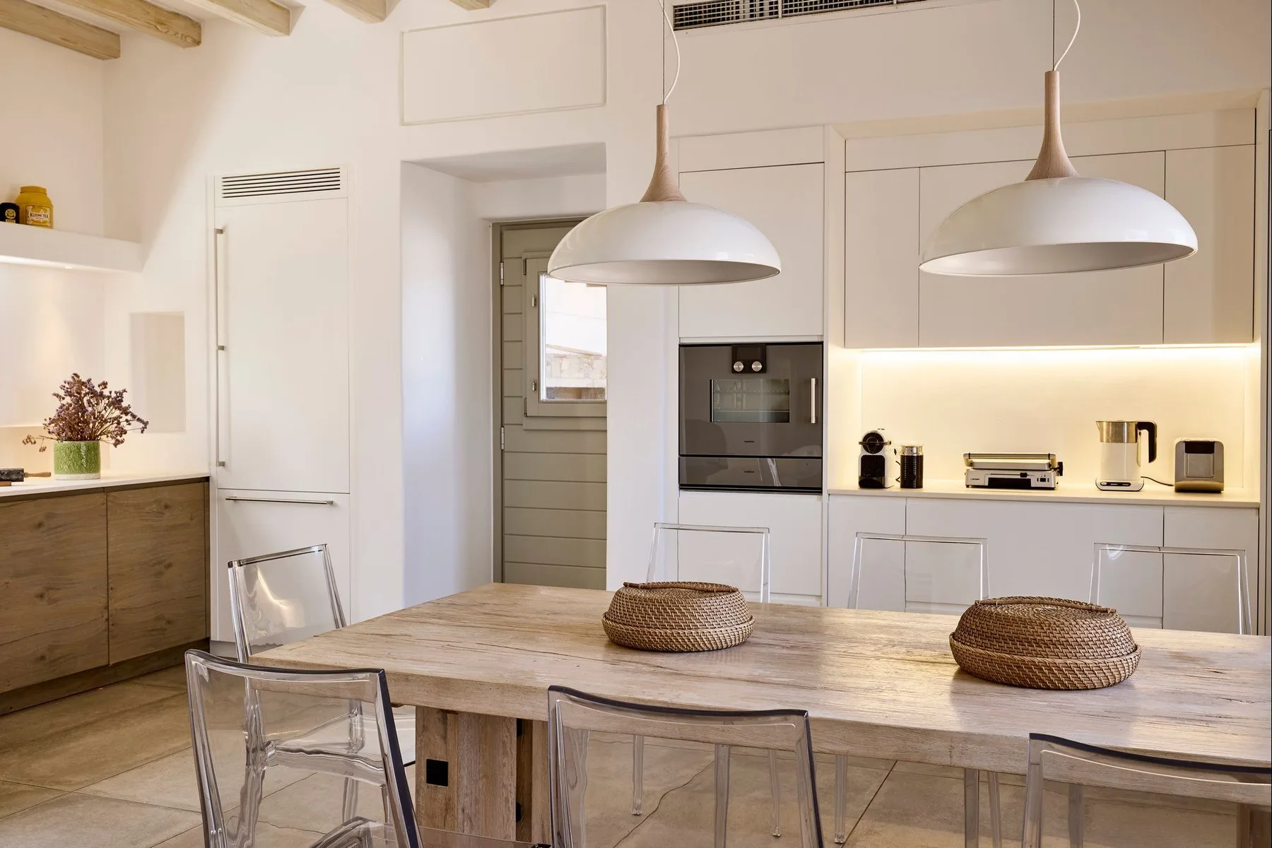 Una cocina luminosa con zona de comedor y una mesa de madera bajo lámparas colgantes.