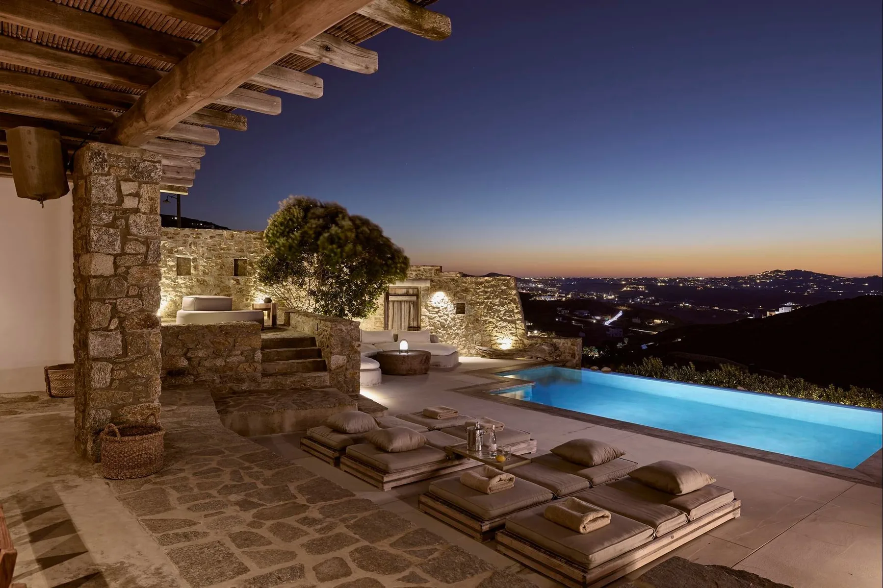 Una terraza de piedra da a una piscina iluminada y colinas lejanas al anochecer.