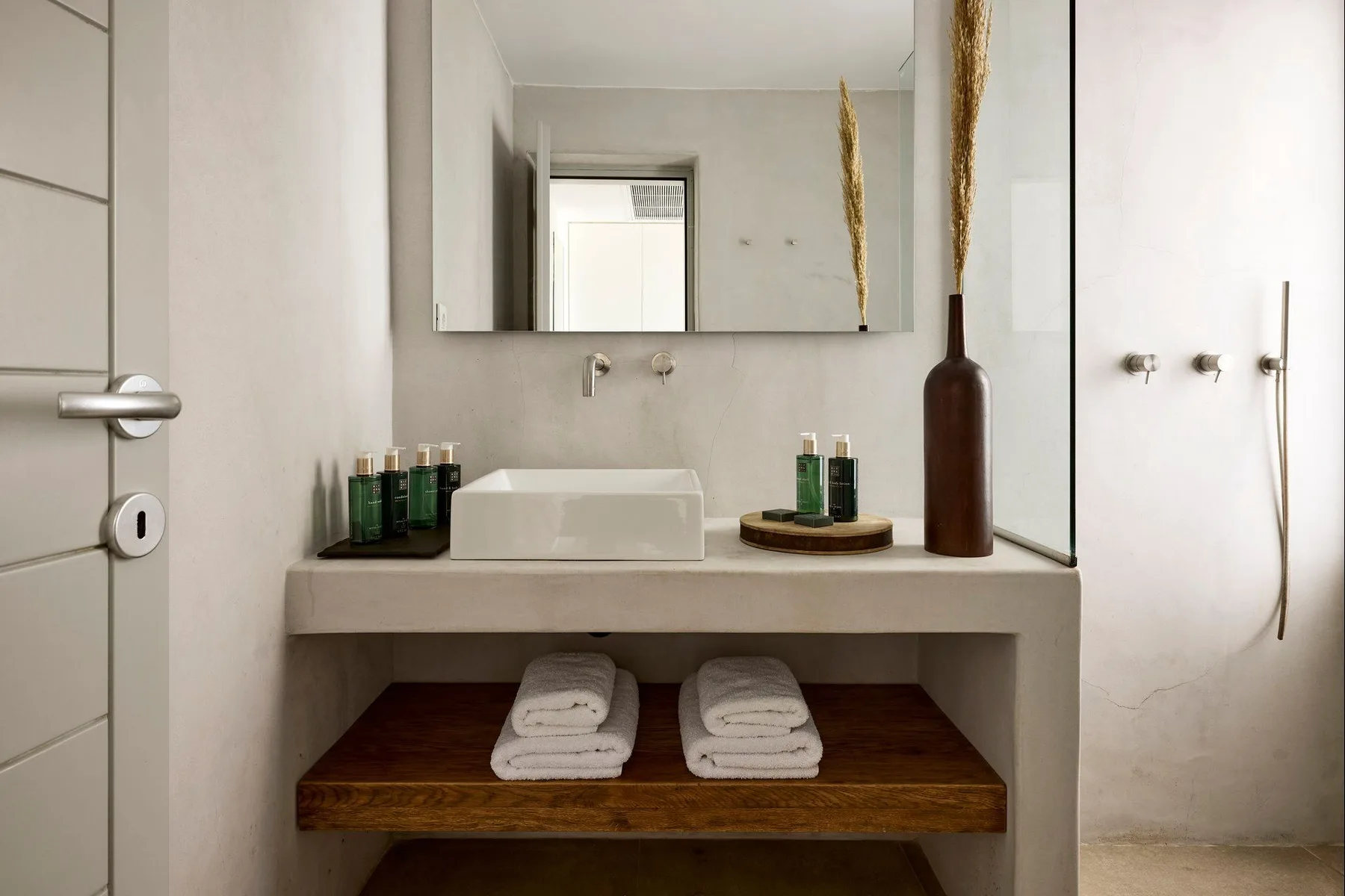 Un tocador de baño con lavabo rectangular, espejo y toallas en un estante abierto.