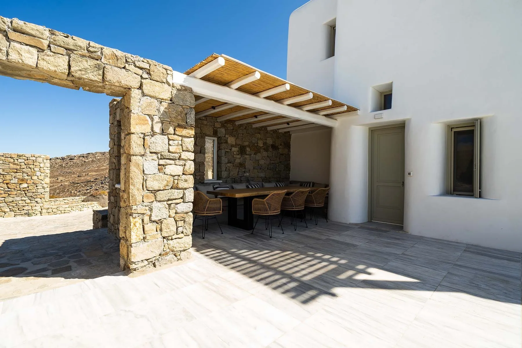 An outdoor dining area sits beneath a pergola beside a white building.
