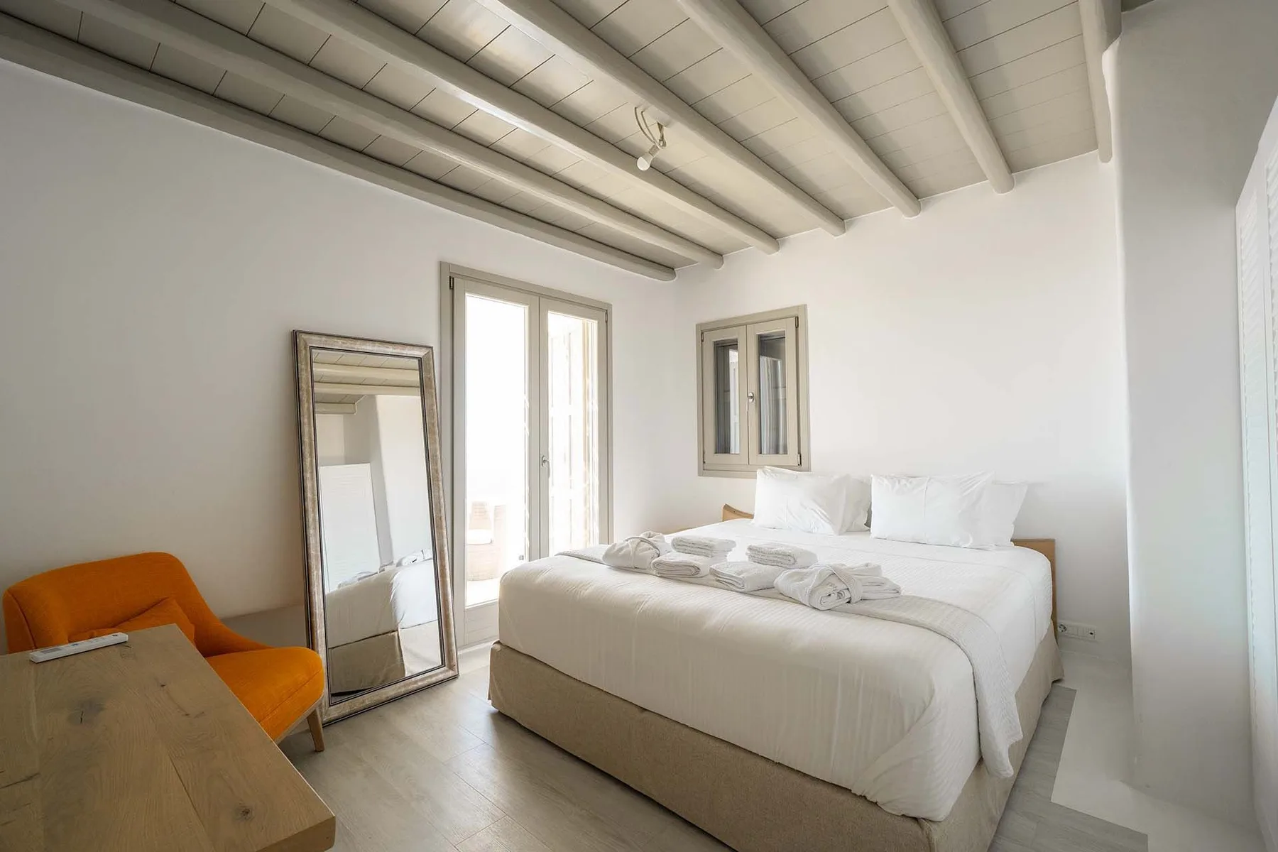 A bright bedroom with a white bed, tall windows, and an orange seat.