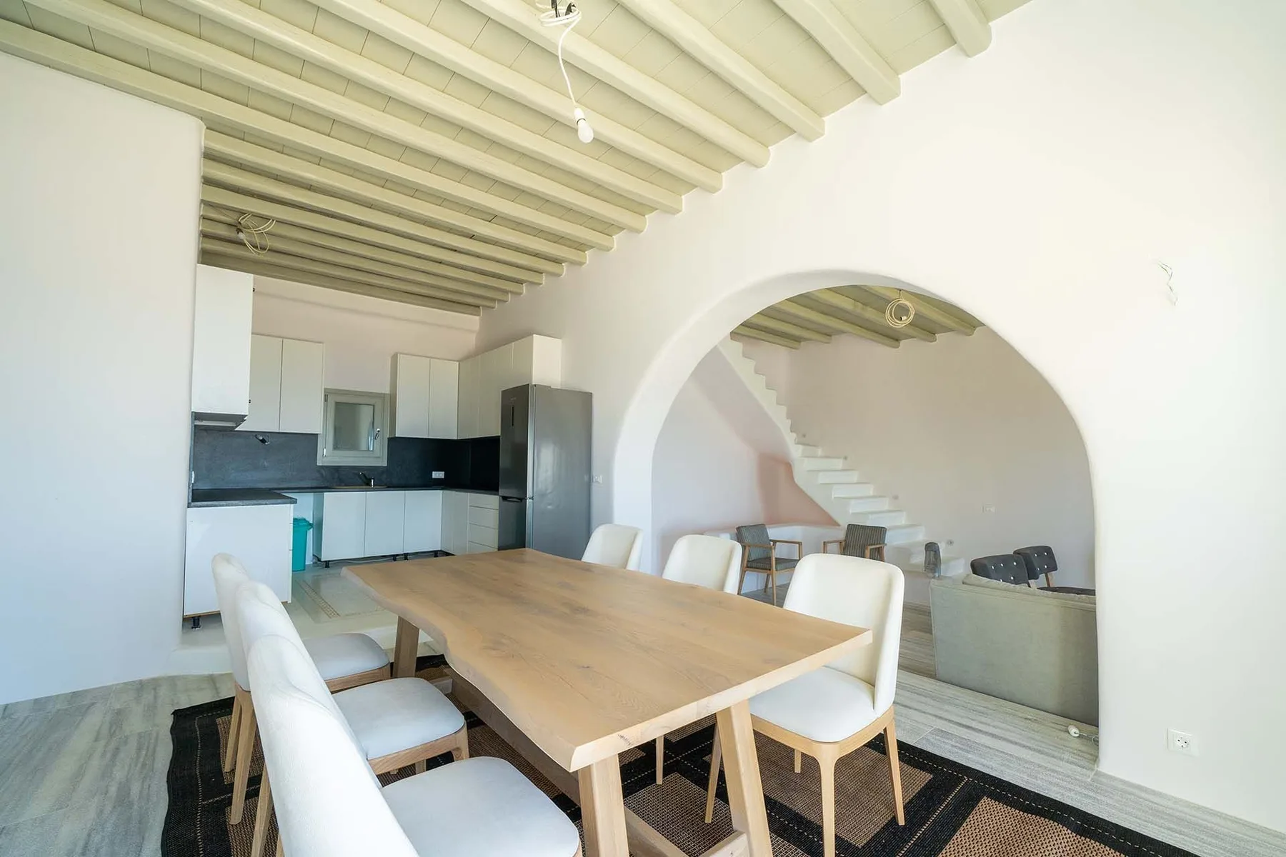 A dining area with a wooden table beside an open kitchen and arched opening.