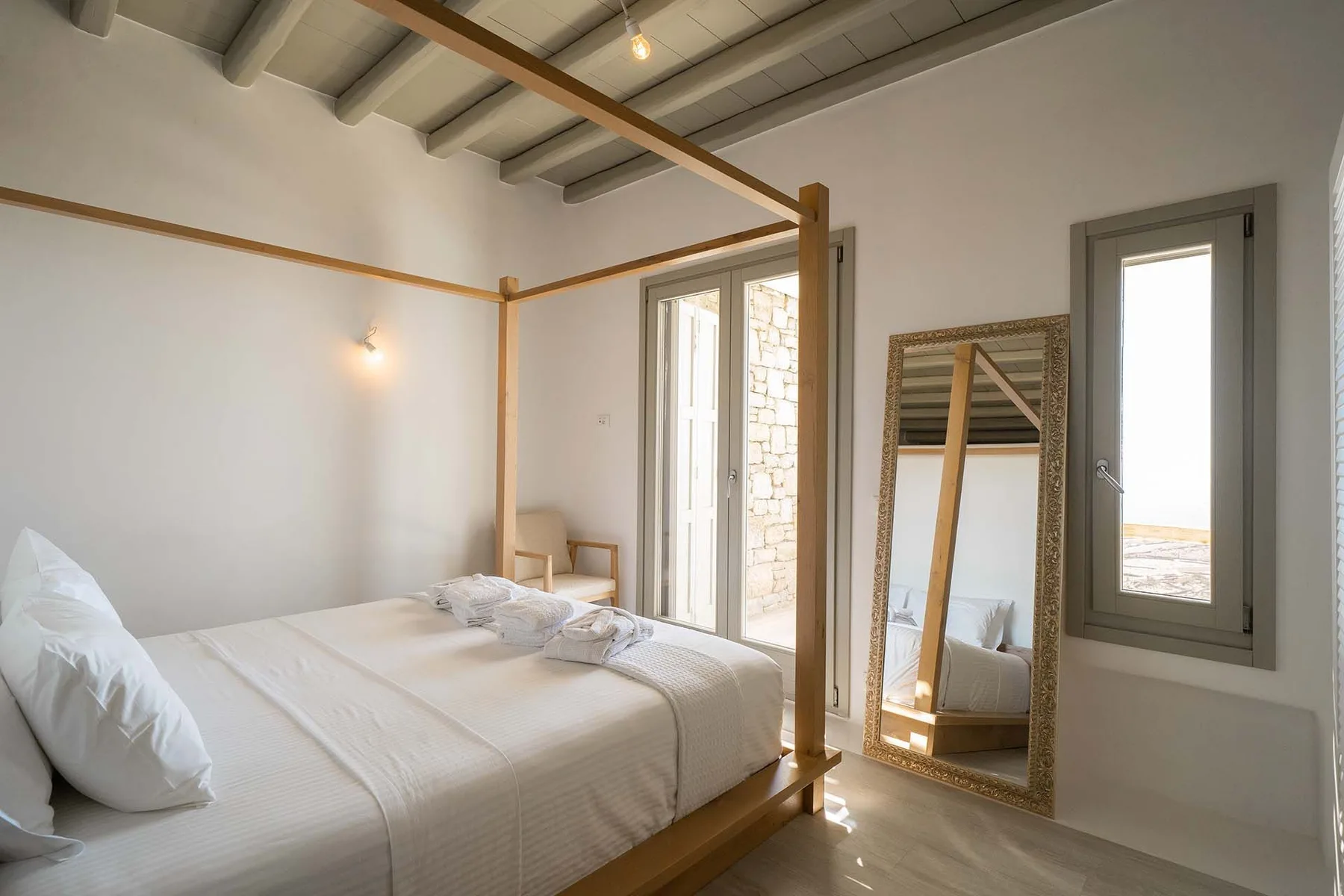 A bright bedroom with a bed, a full-length mirror, and tall windows.