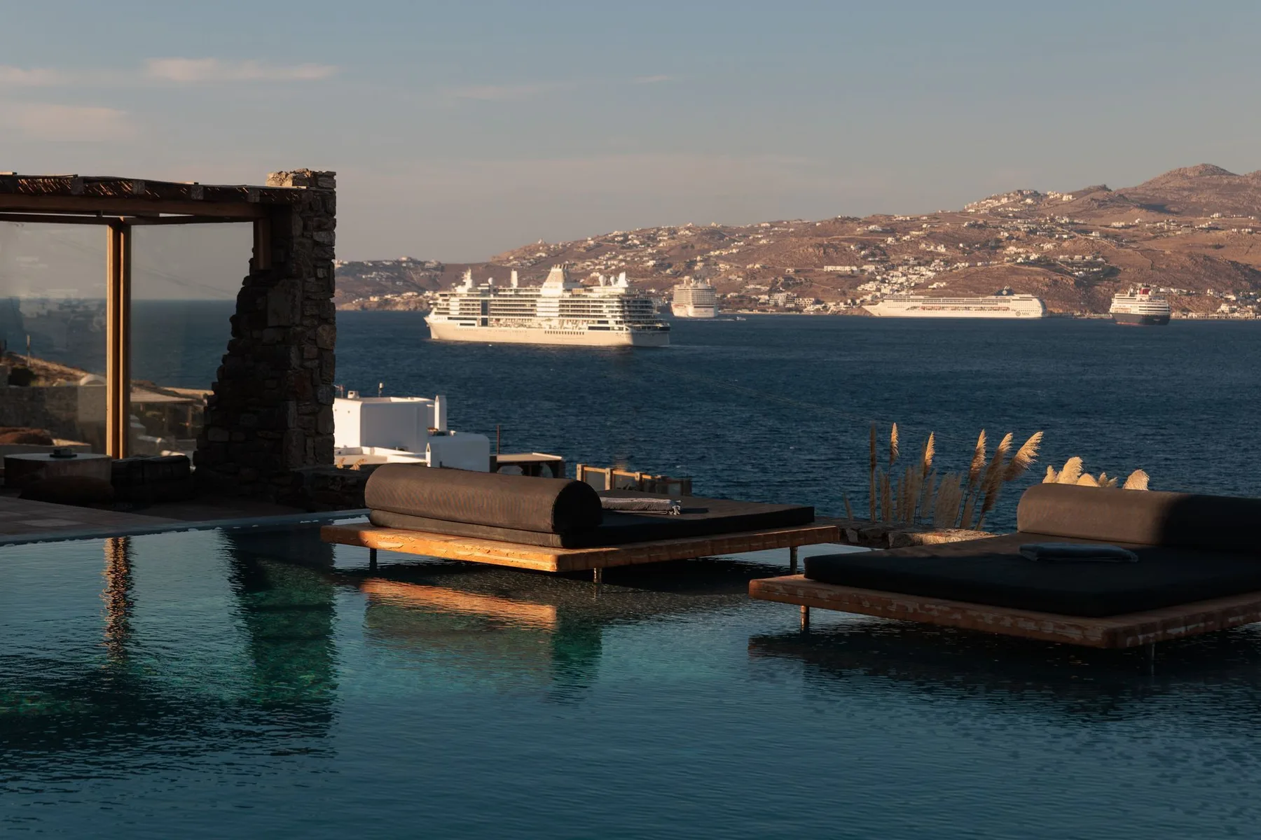 An infinity pool overlooks the sea, with cushioned loungers and cruise ships beyond.