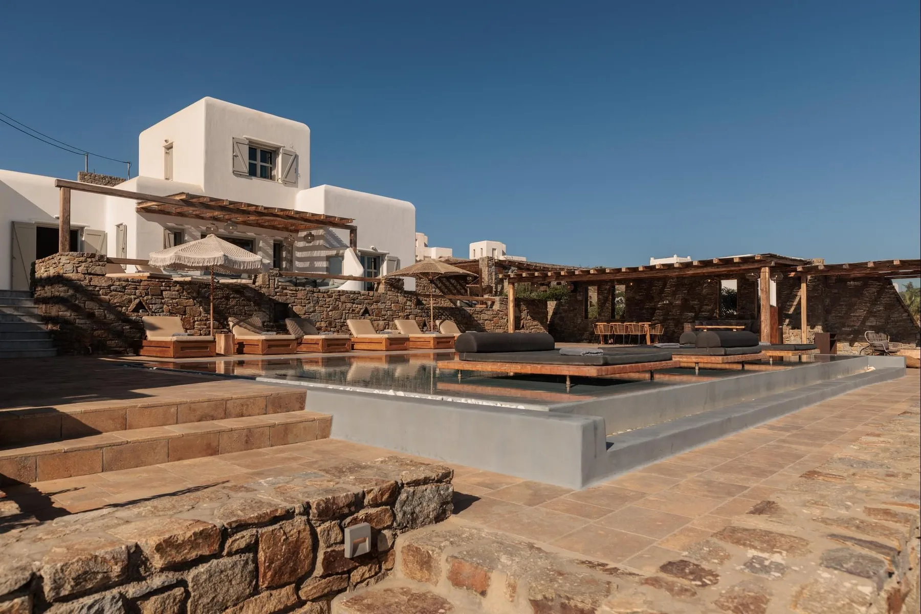 Outdoor pool beside a stone terrace and white buildings under a clear sky.