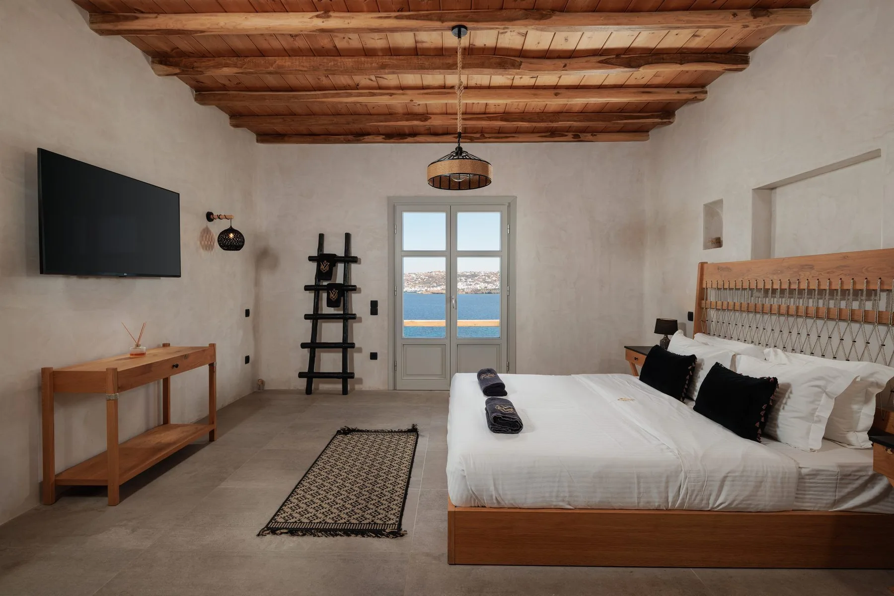 A bedroom with a large bed, wooden ceiling beams, and doors opening to a sea view.