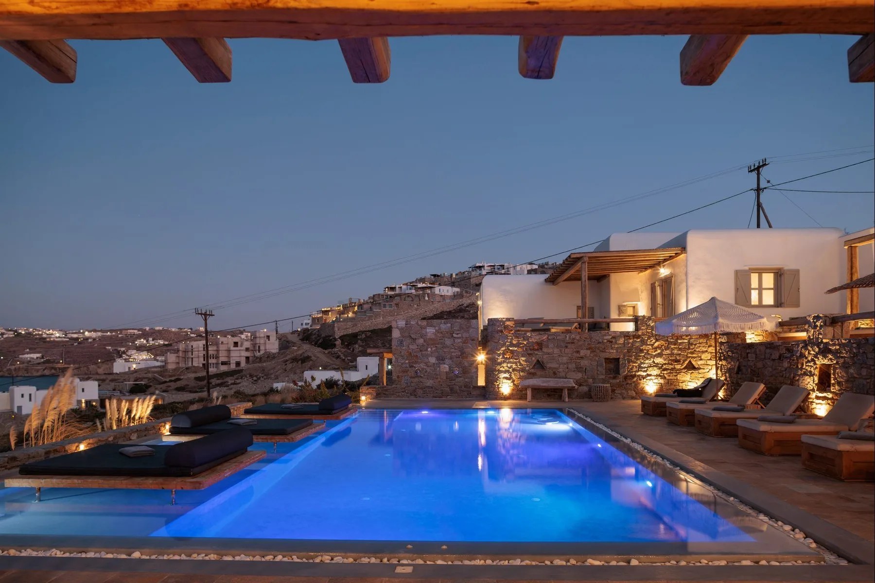 An outdoor pool beside a stone terrace and illuminated white buildings at dusk.