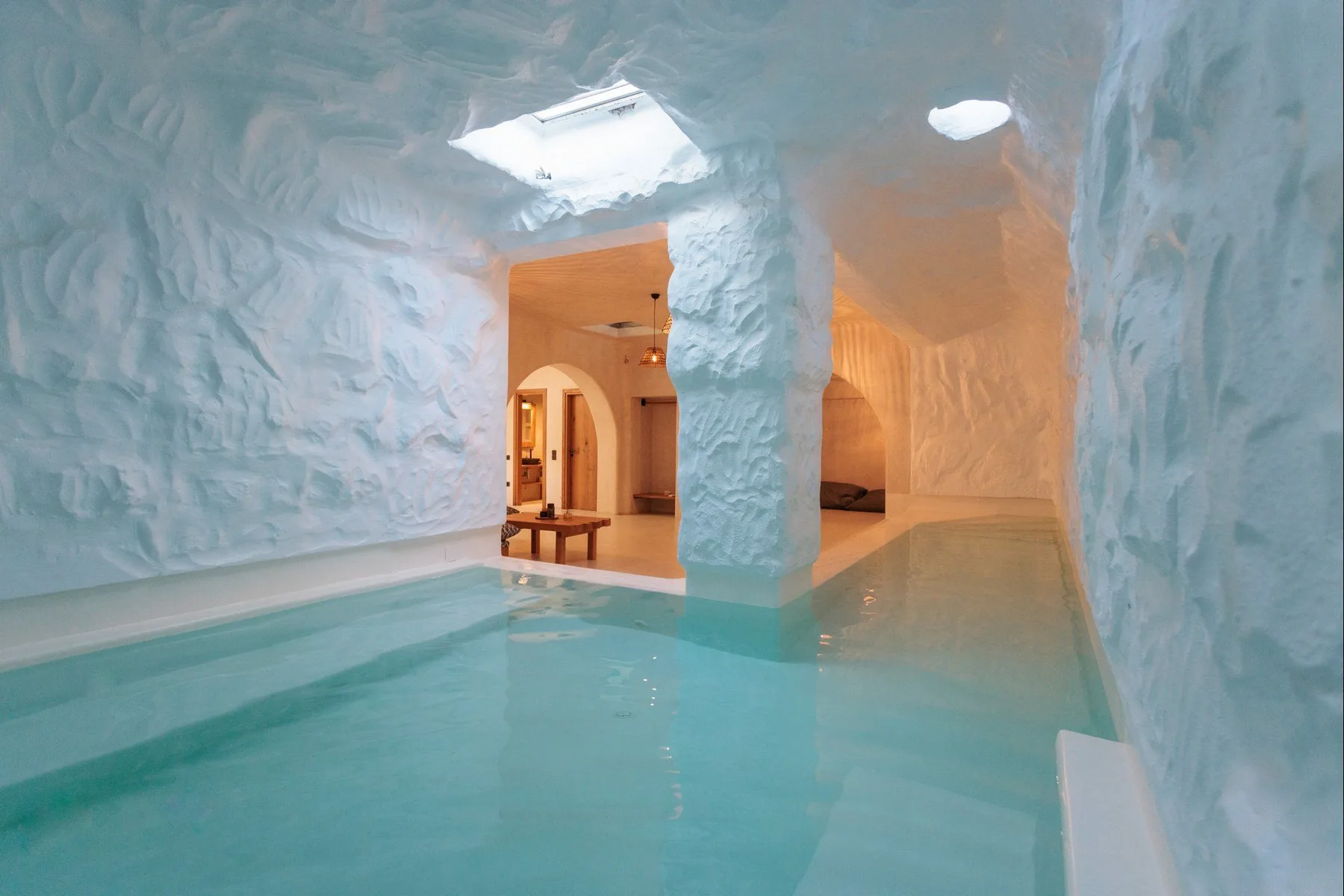 An indoor pool sits within sculpted pale walls, with warm-lit openings beyond.