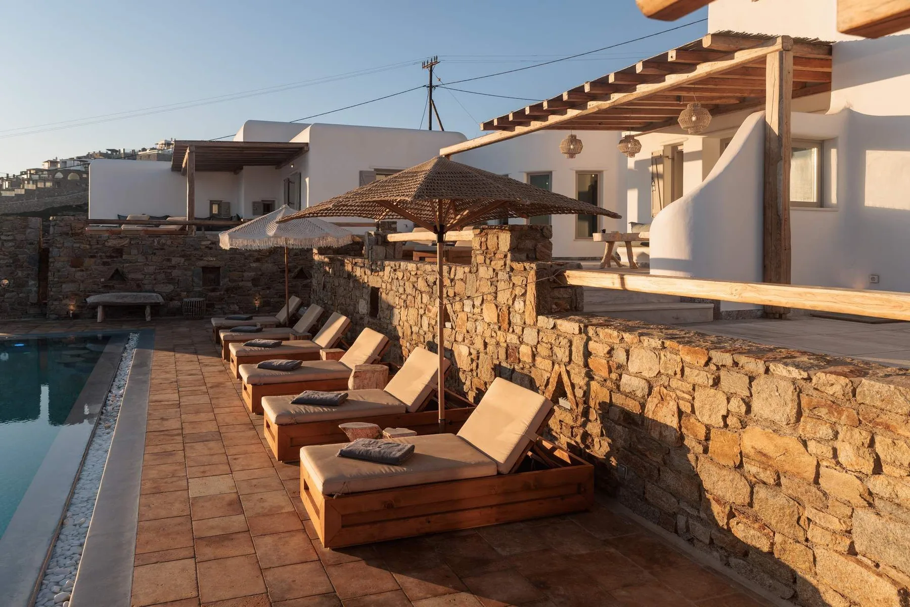 A poolside patio with wooden loungers, stone walls, and a shaded pergola.