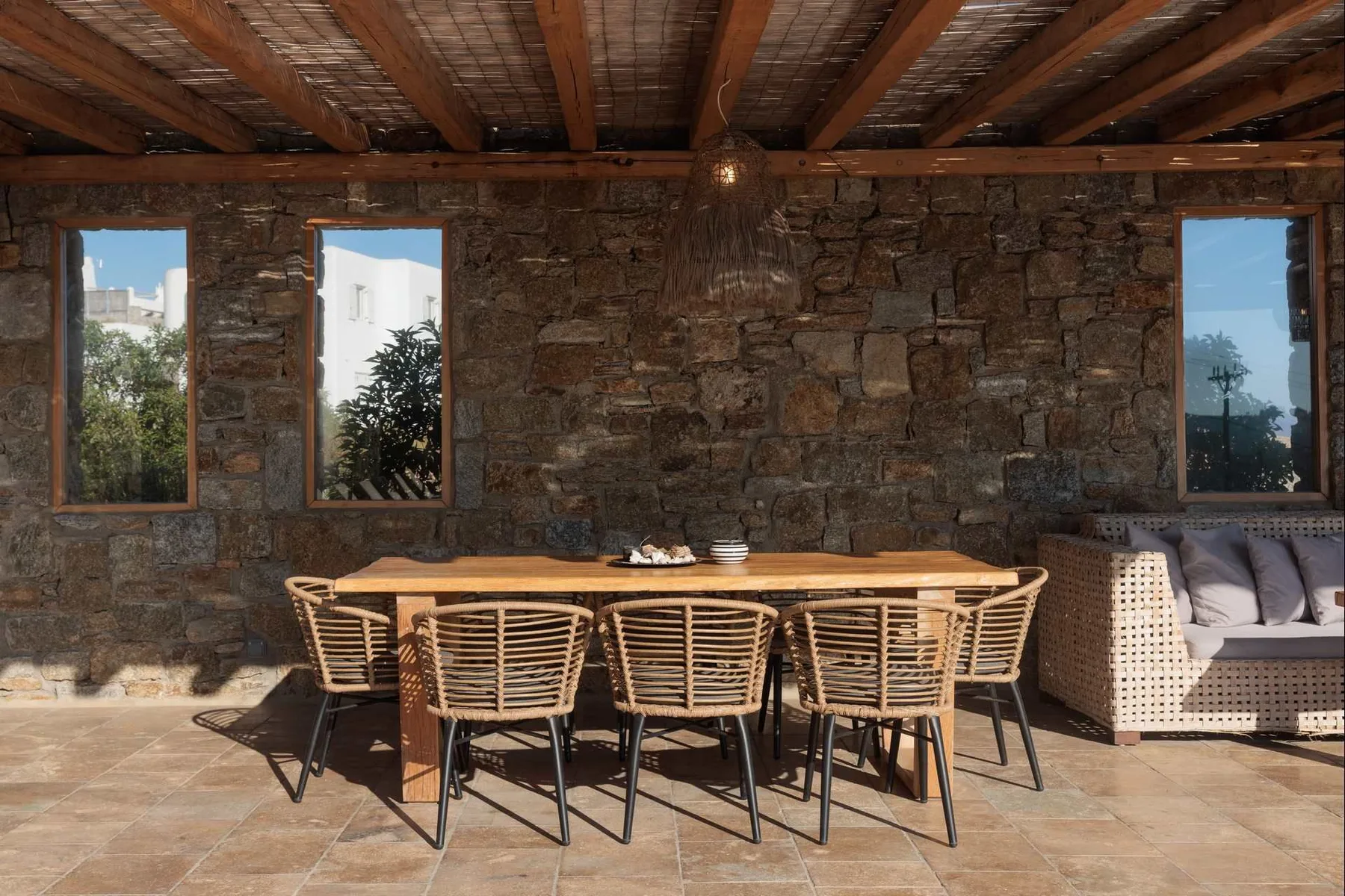A rustic dining area with a long table and woven seating beneath exposed wooden beams.