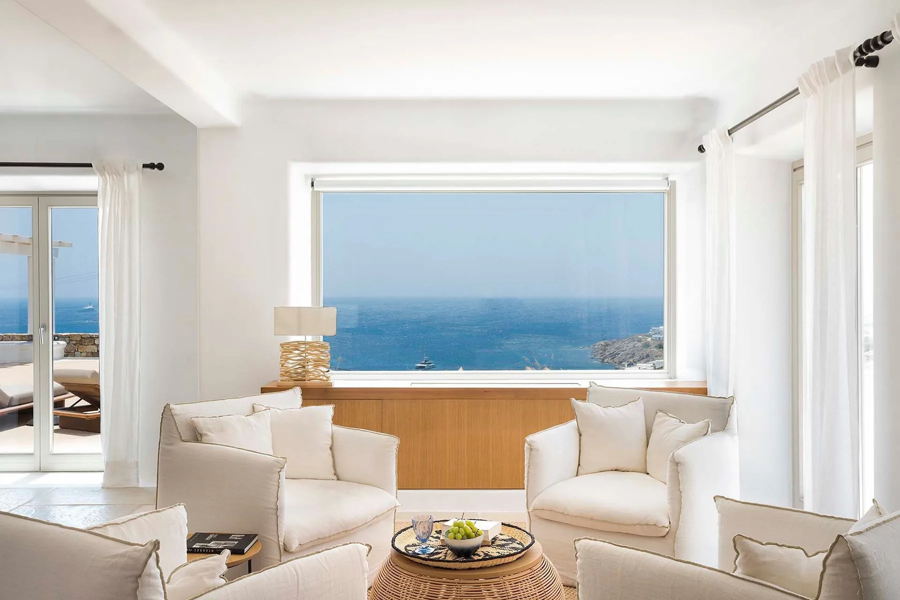 A bright living room with white seating and a large window overlooking the sea.