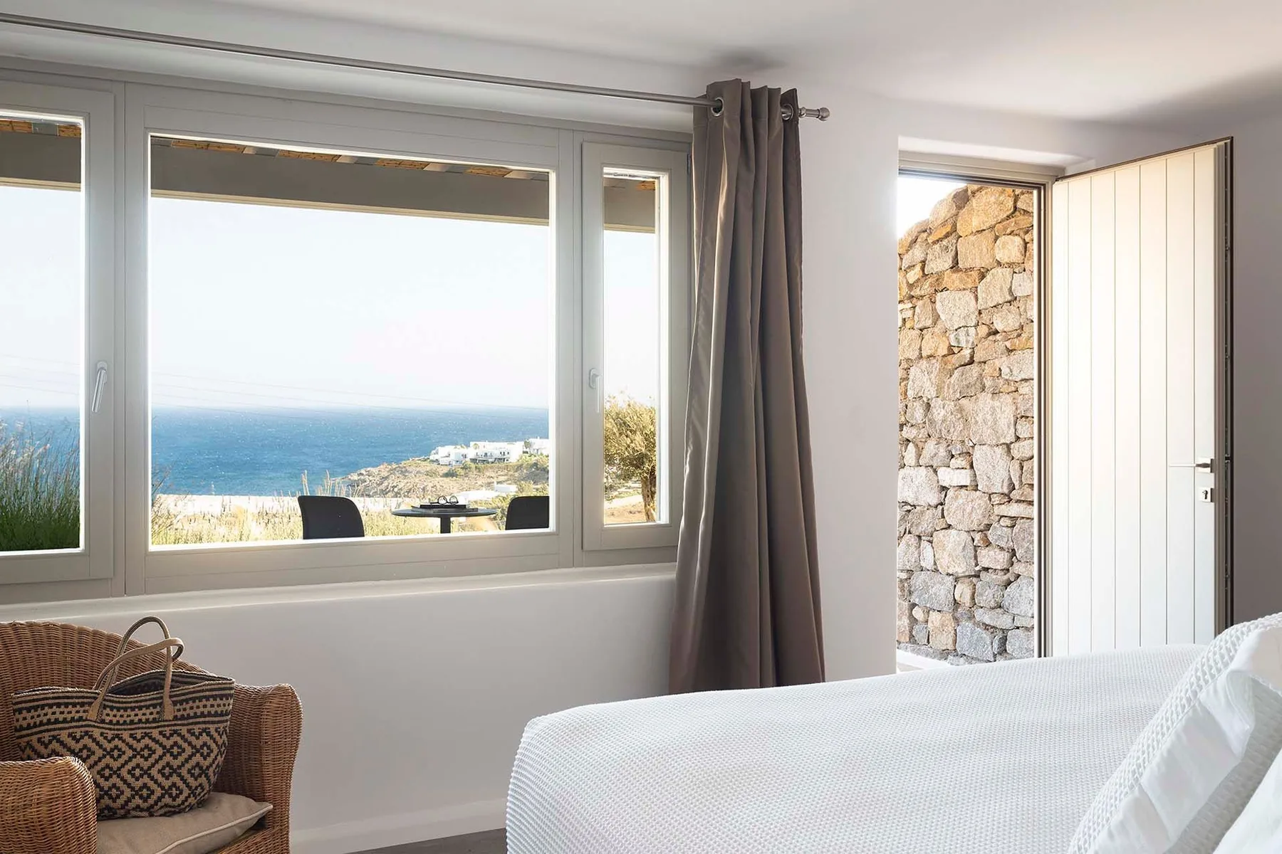 A bright bedroom overlooks the sea through large windows, with a bed beside an open doorway.