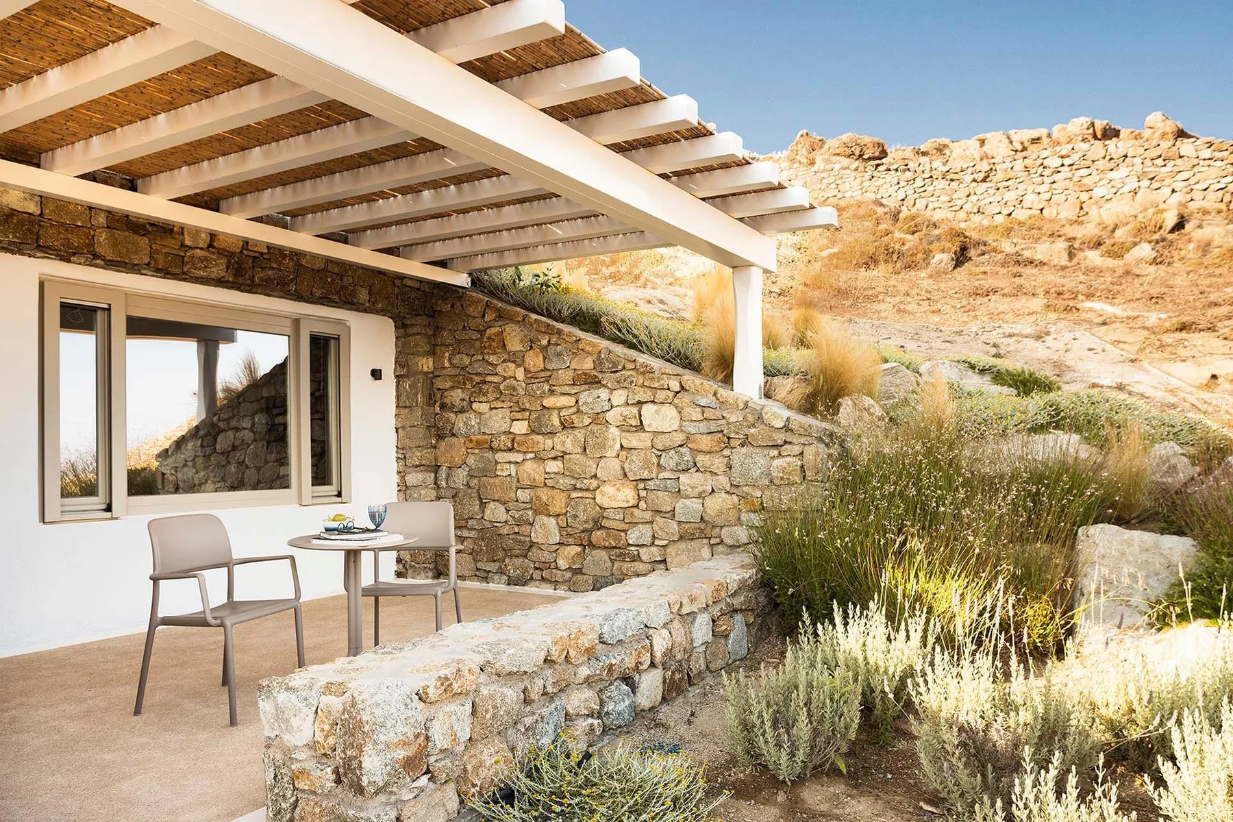 A stone patio beneath a white pergola overlooks dry, rocky hills.