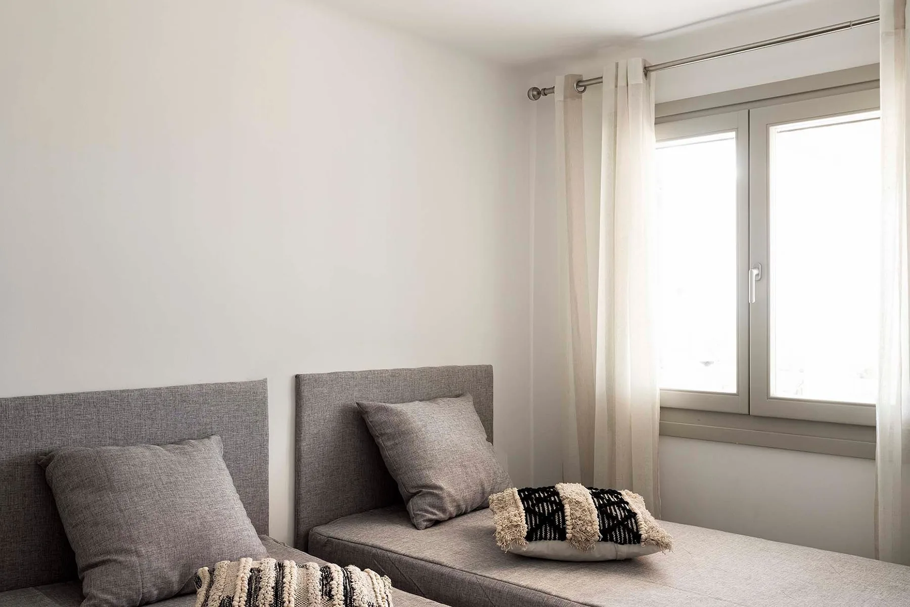 A bedroom with two gray beds beside a window with light curtains.