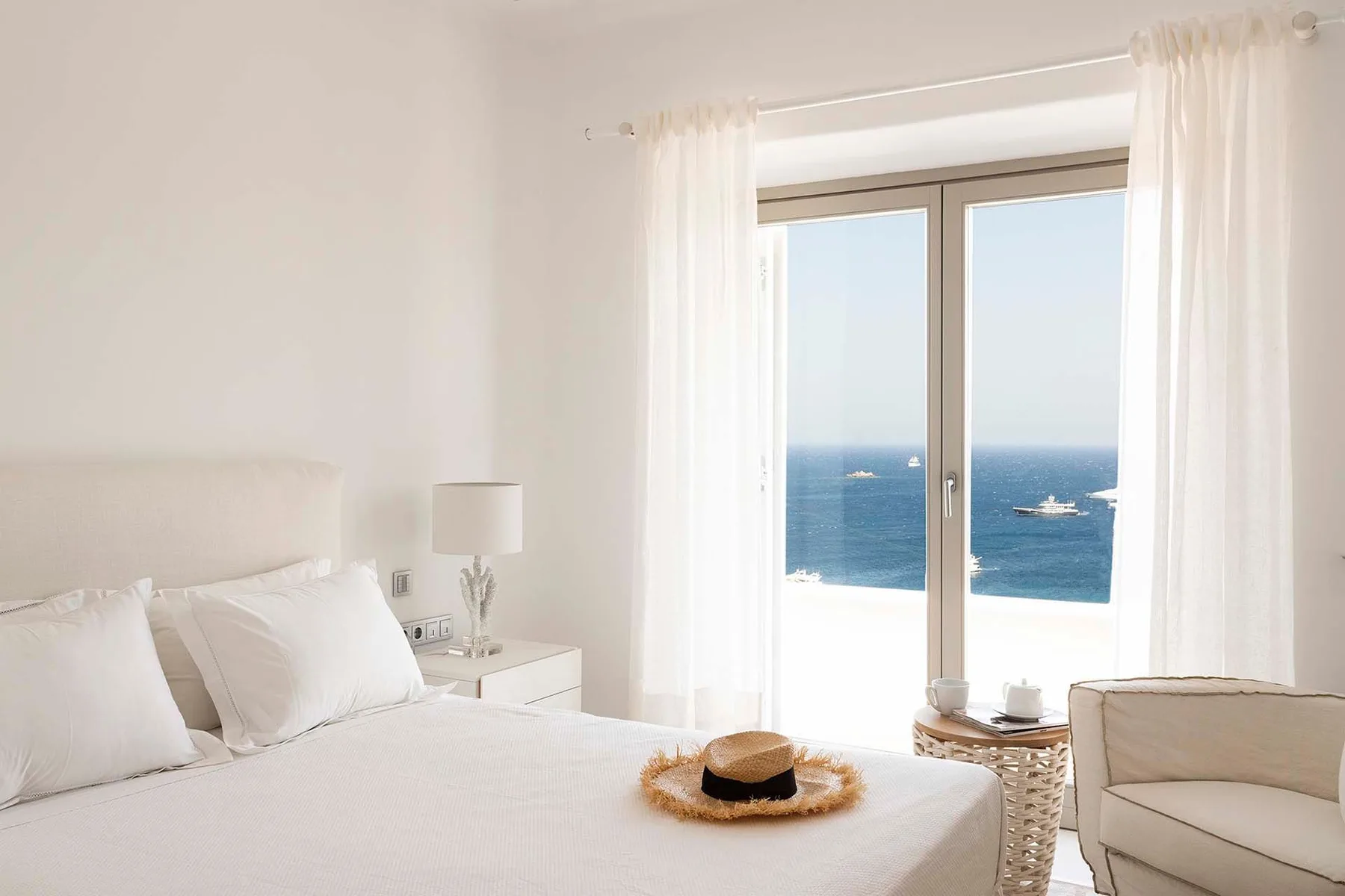 A bright bedroom with a bed beside a large window overlooking the sea.