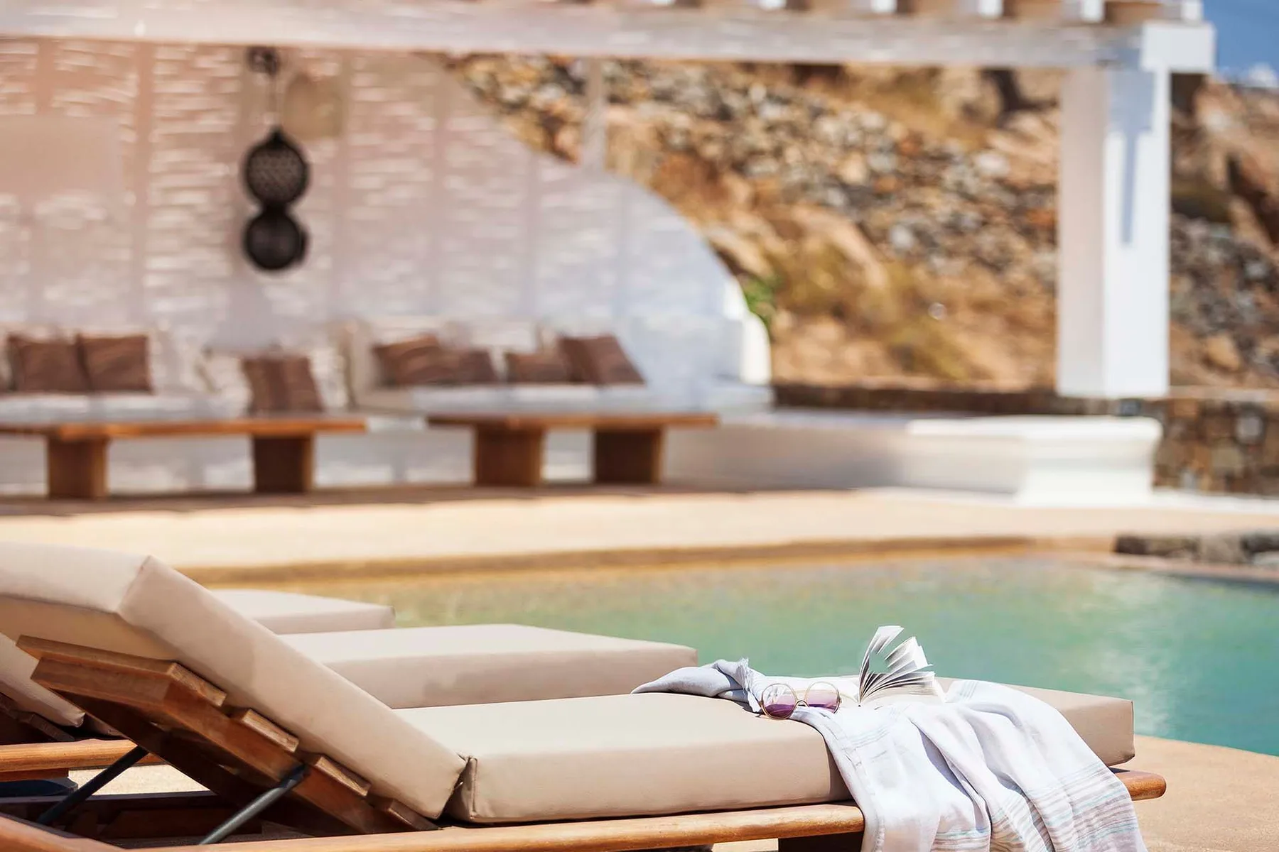 Poolside loungers sit beside a swimming pool, with a shaded seating area behind them.