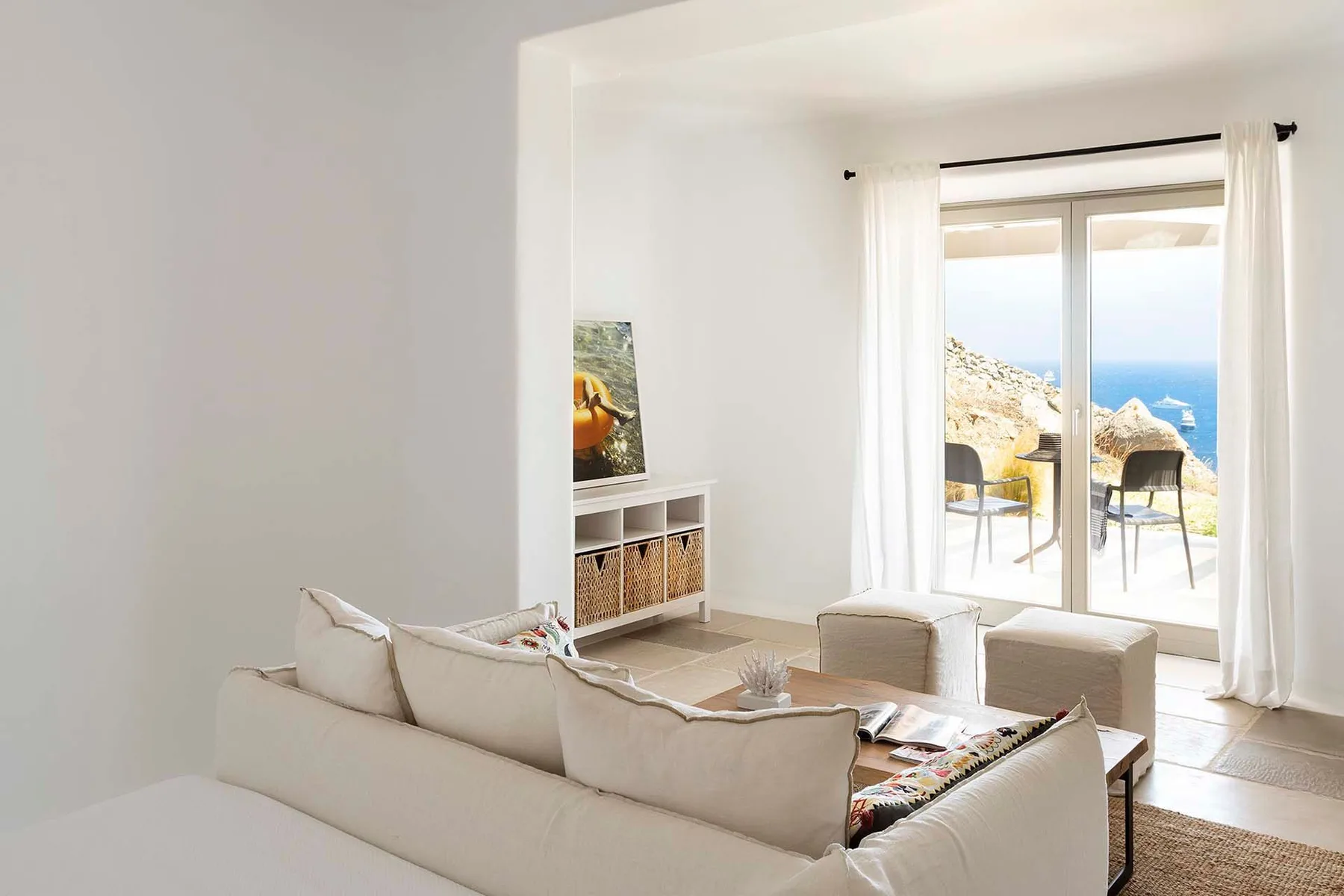 A bright living room with pale seating and glass doors opening onto a sea view.