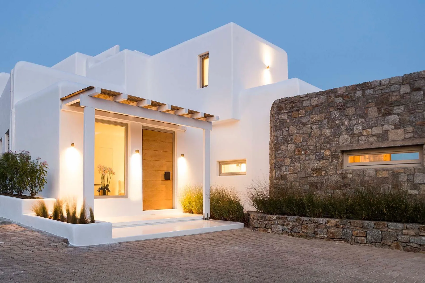 A modern white building entrance with a wooden door and stone wall at dusk.