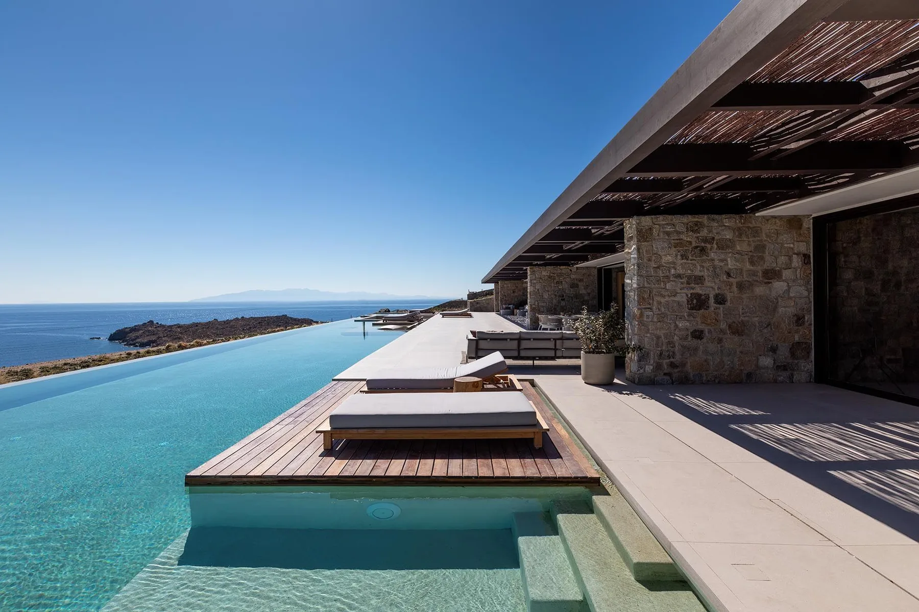 Una piscina infinita al aire libre junto a una terraza de piedra con sombra da al mar.