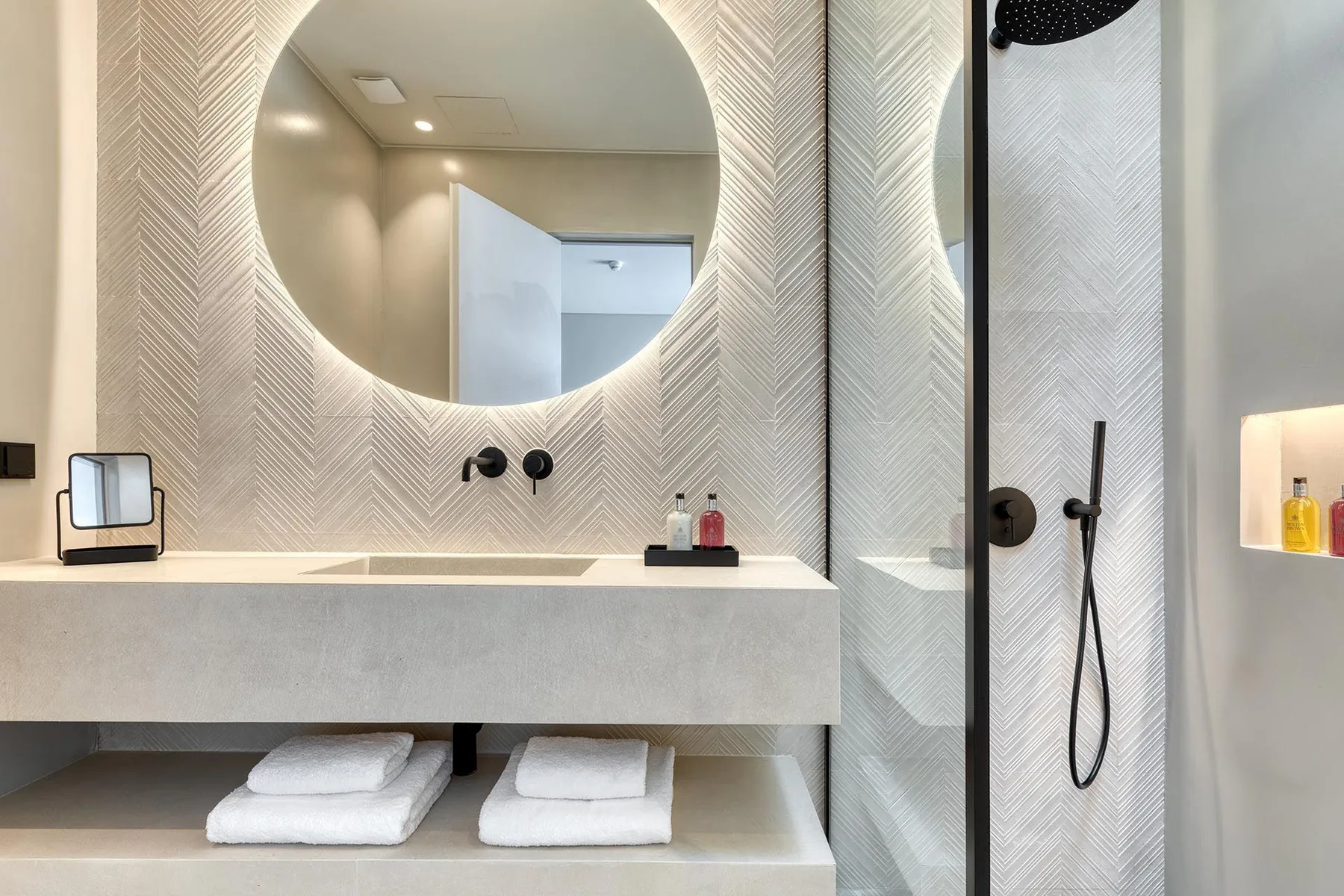 Un baño luminoso con un amplio lavabo, espejo redondo y ducha de cristal.
