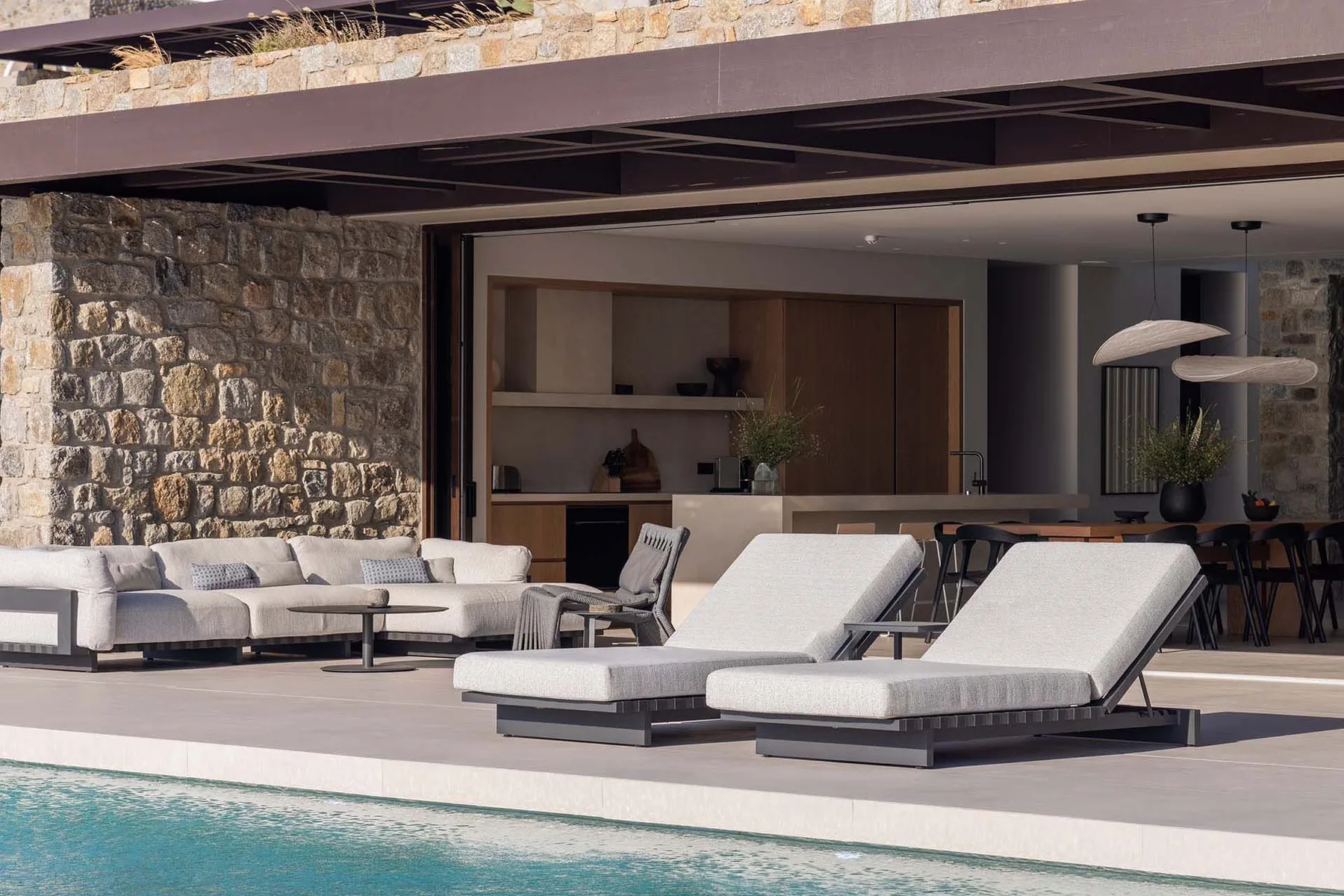 Un patio junto a la piscina con tumbonas acolchadas y asientos bajo una zona cubierta.