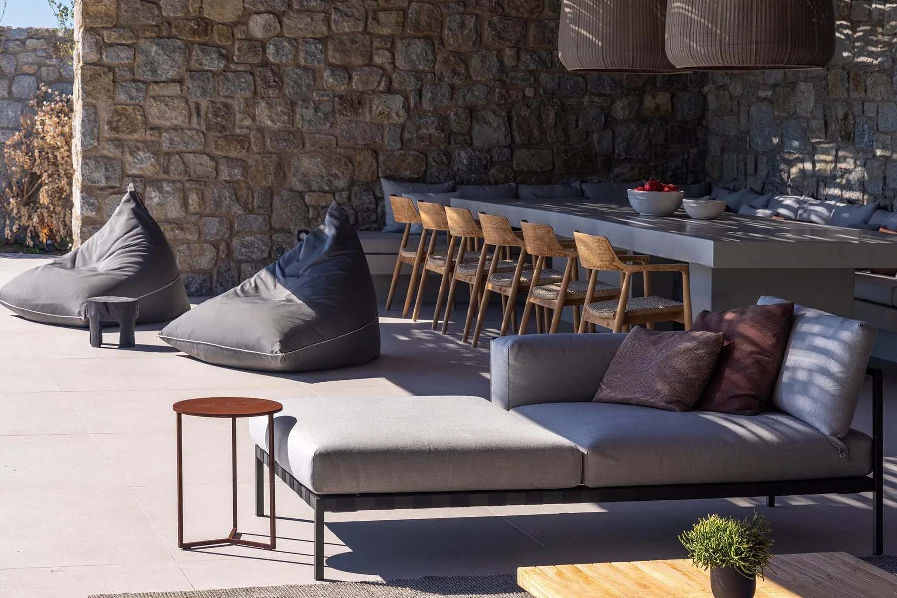 Una sala exterior con asientos acolchados y una mesa larga junto a un muro de piedra.