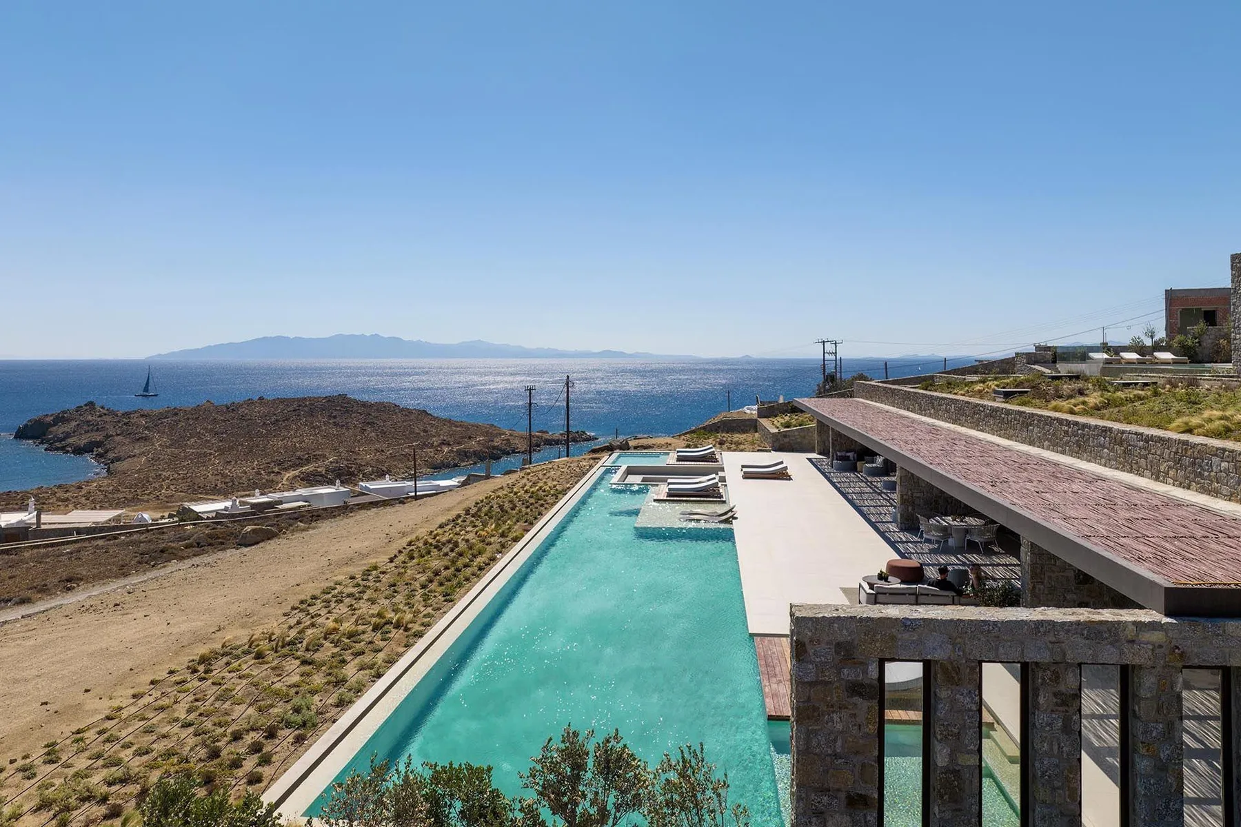Una larga piscina al aire libre mira al mar junto a una terraza soleada.