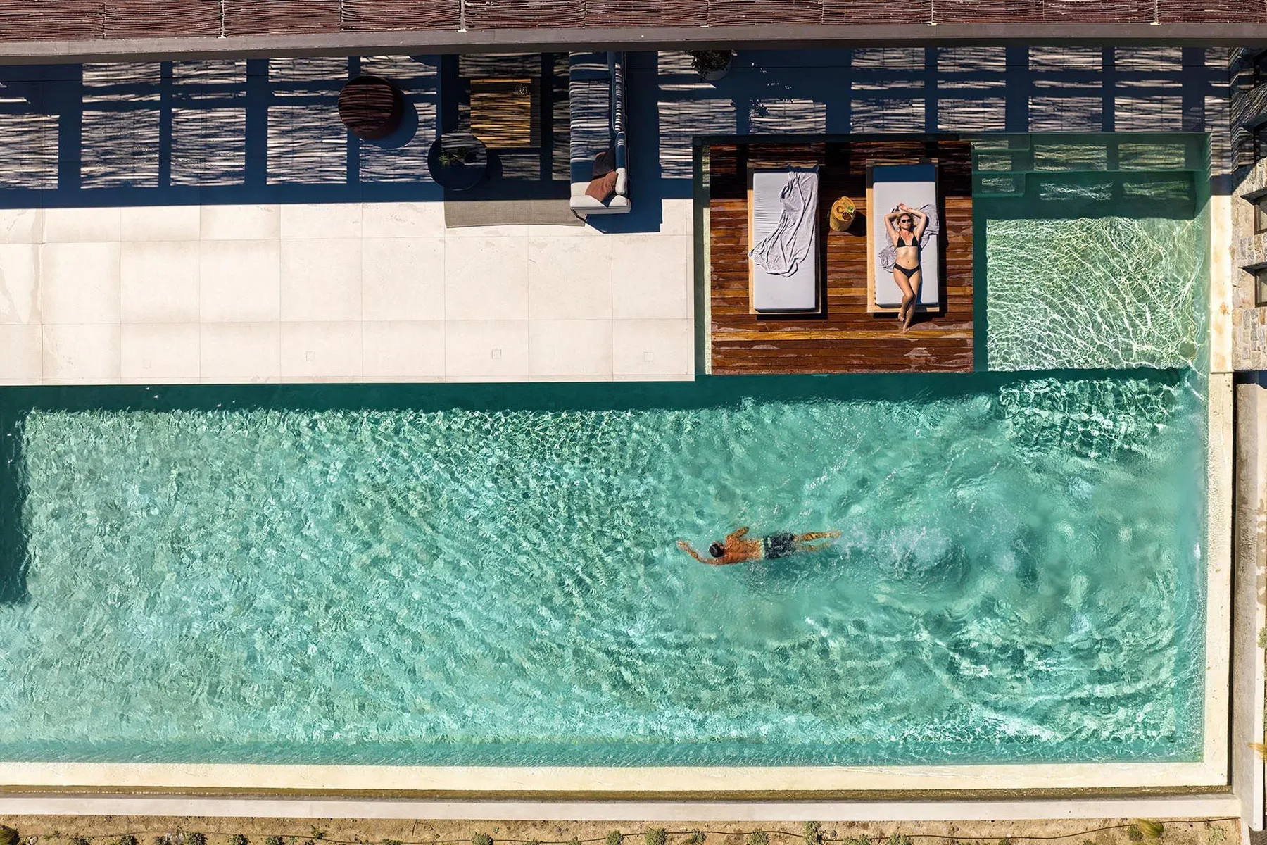 Una piscina al aire libre con una persona nadando y gente descansando en la terraza.