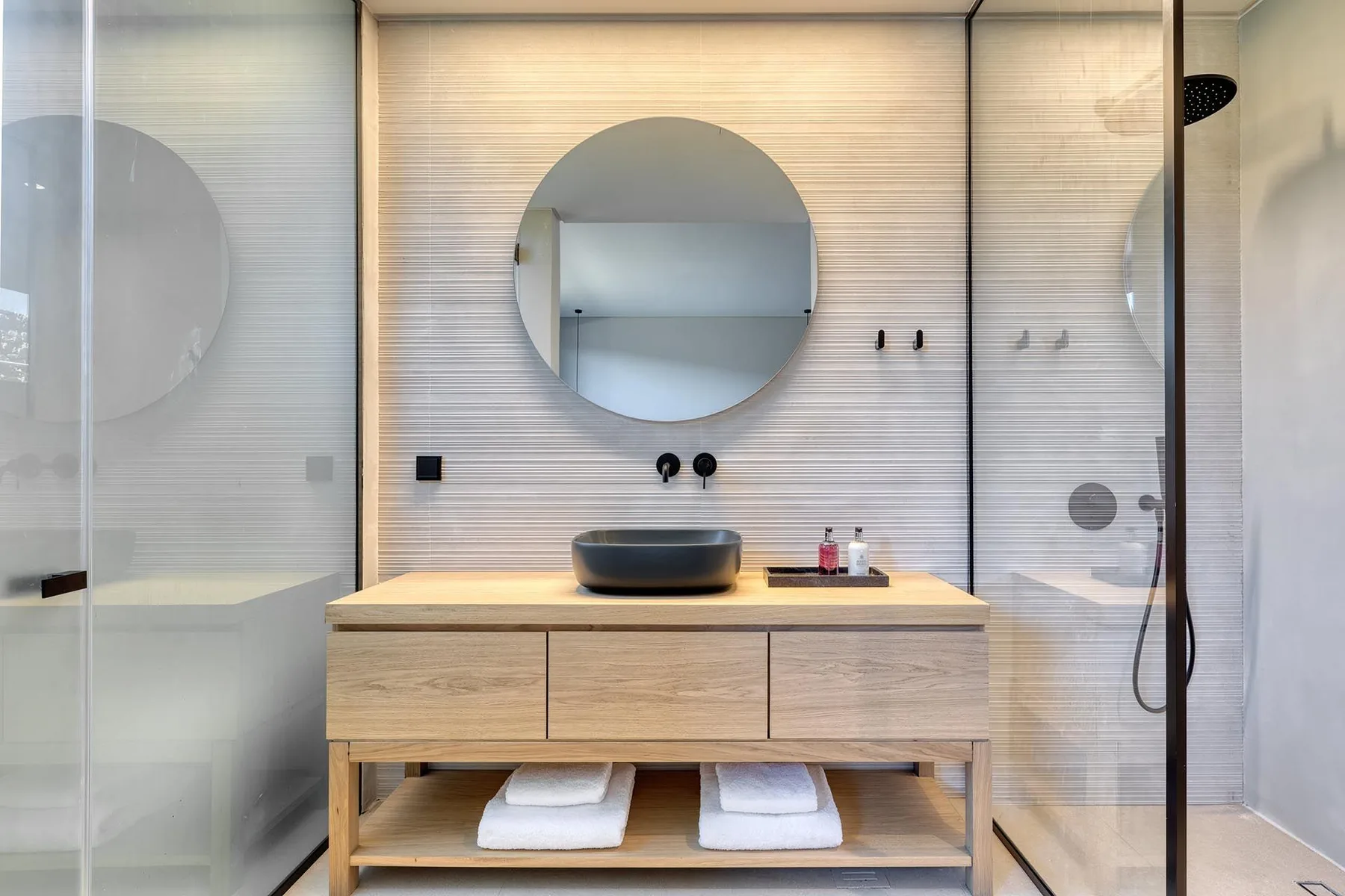 Un baño moderno con un mueble de madera, espejo redondo y ducha de vidrio.