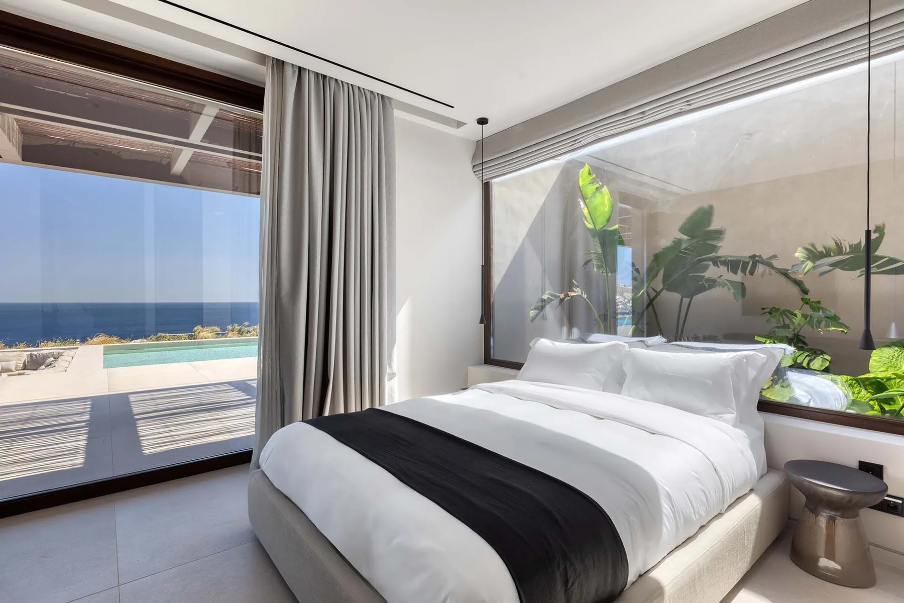 Un dormitorio luminoso con una cama bien tendida junto a una gran ventana con vistas al mar.