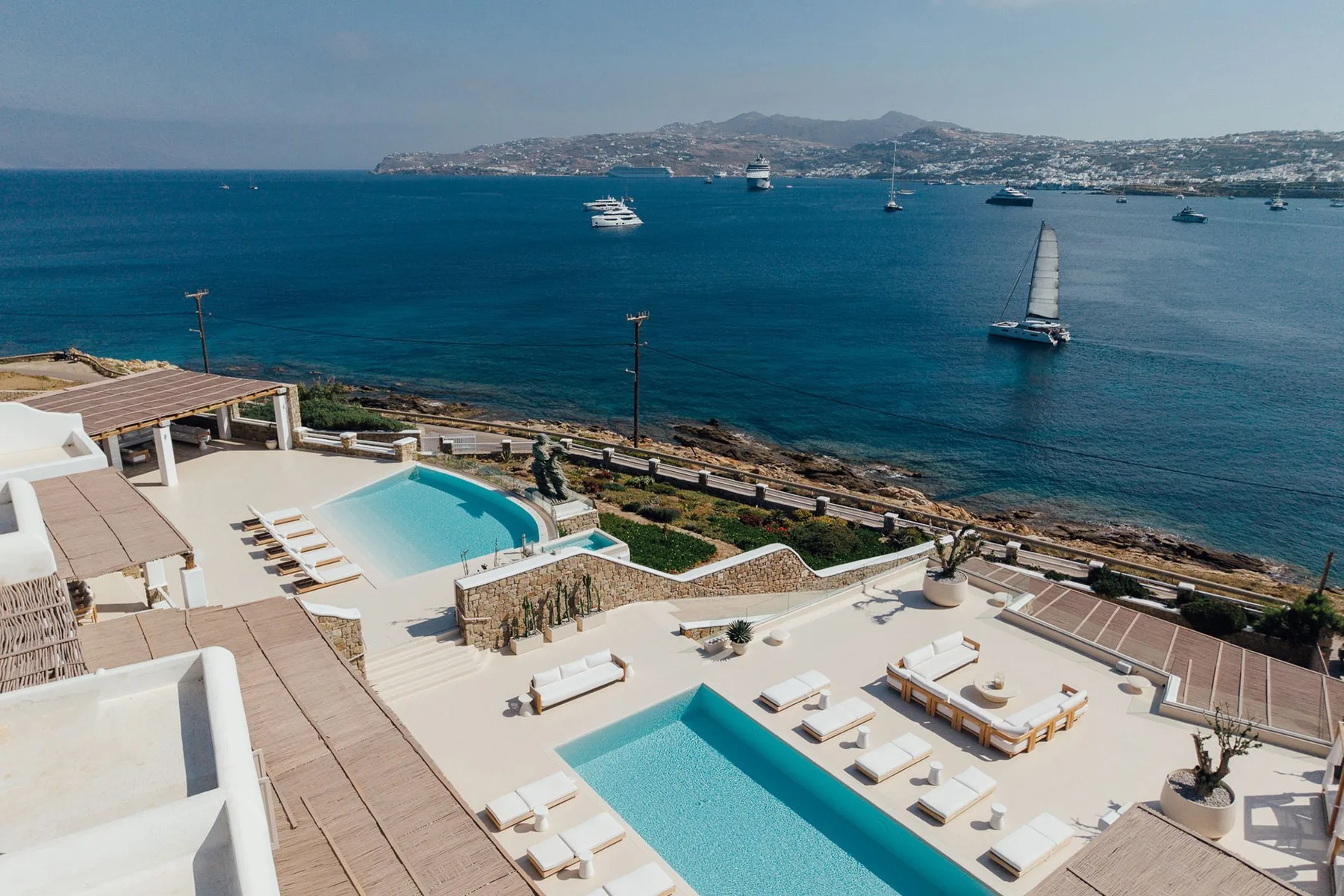 A seaside terrace with swimming pools overlooks a bay dotted with sailboats.