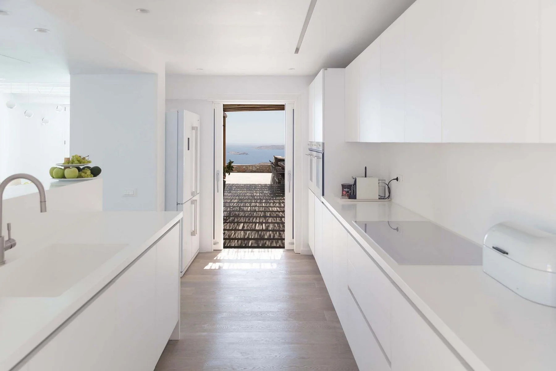 A bright white kitchen with long counters and a view of the sea through the doorway.