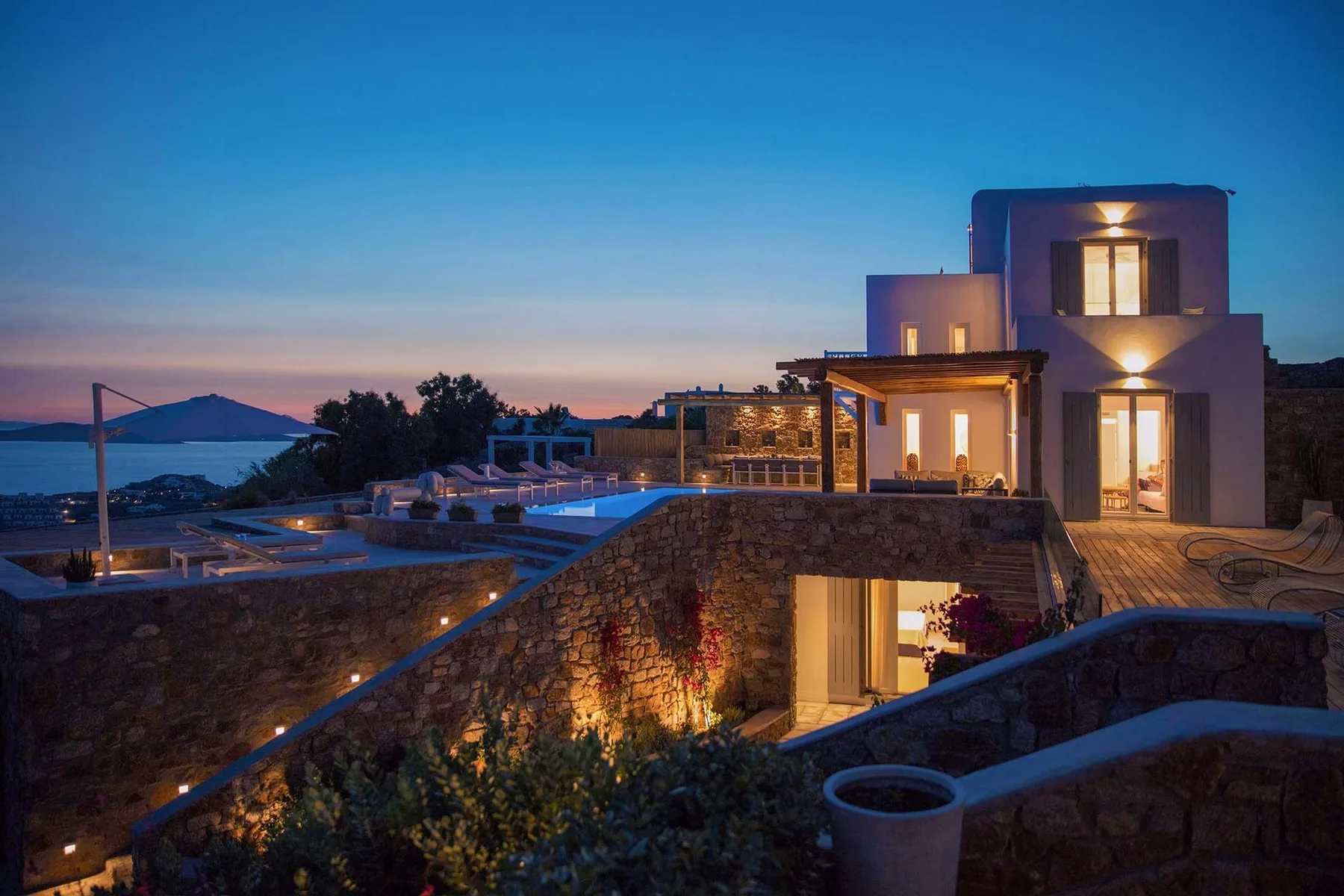 A modern home glows at dusk beside a swimming pool overlooking the sea.