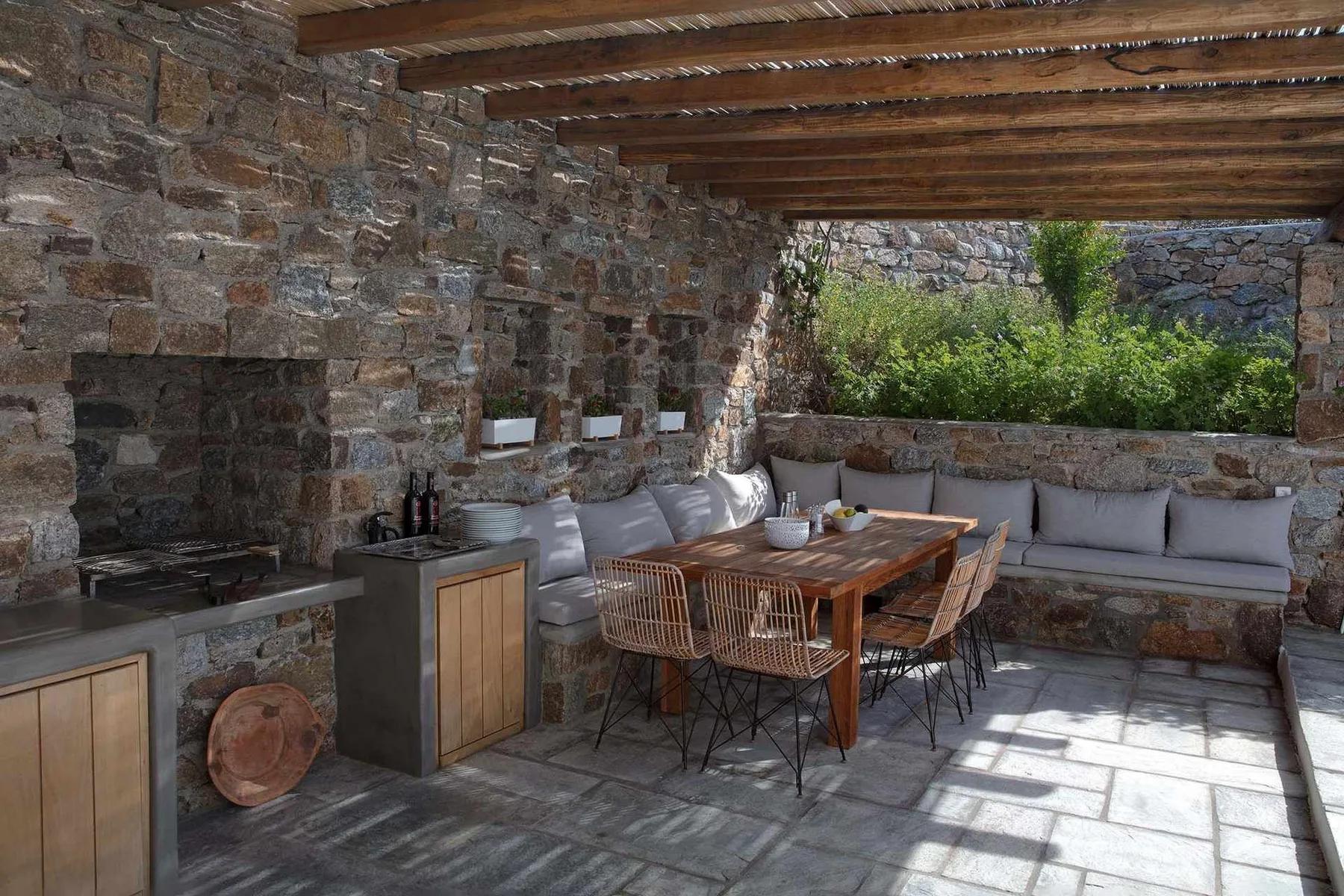 A covered stone patio has a wooden dining table and built-in seating.