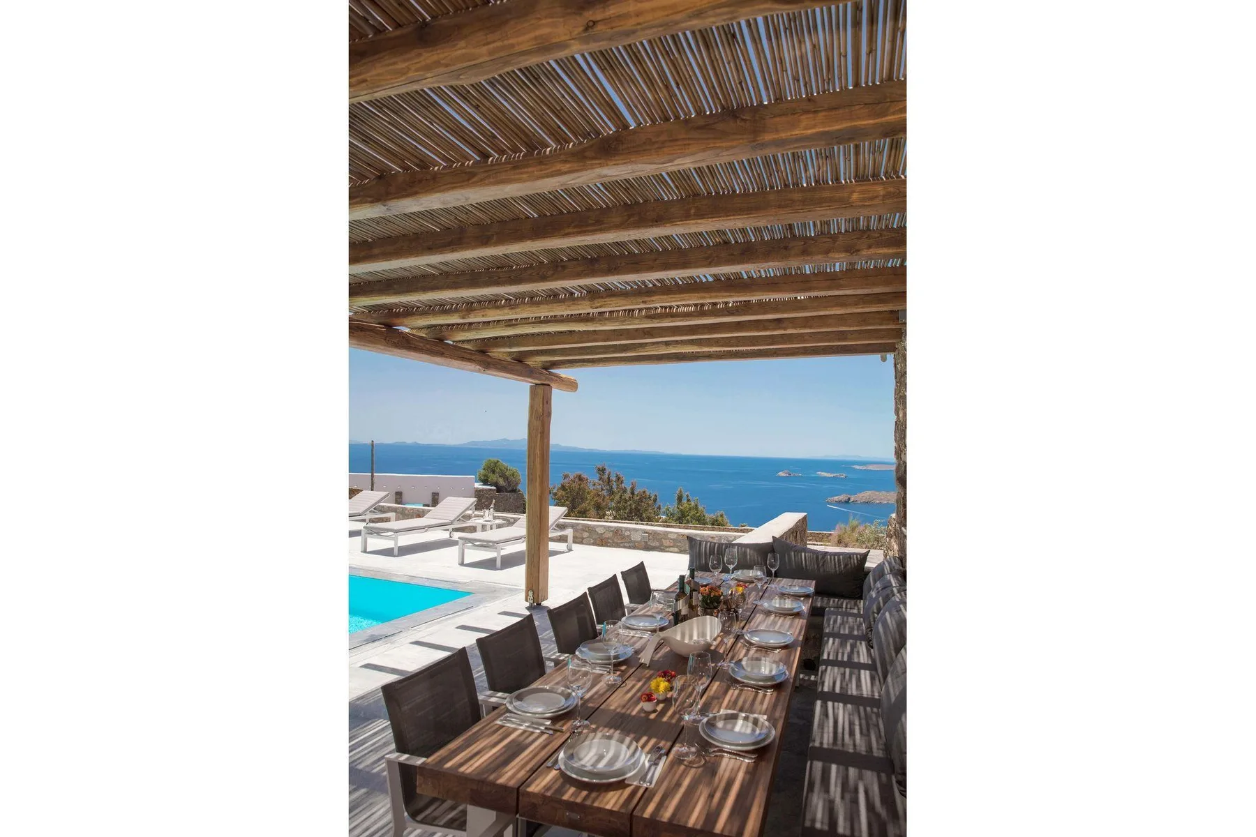 An outdoor dining area under a wooden pergola overlooks a pool and the sea.