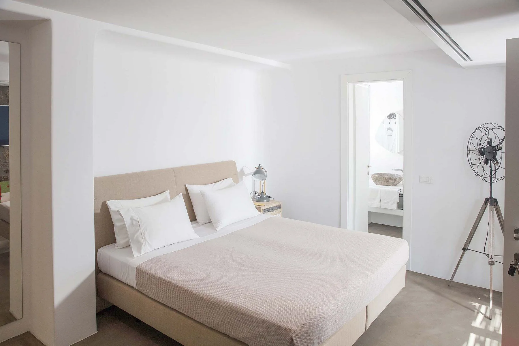A bedroom with a neatly made bed and a standing fan.