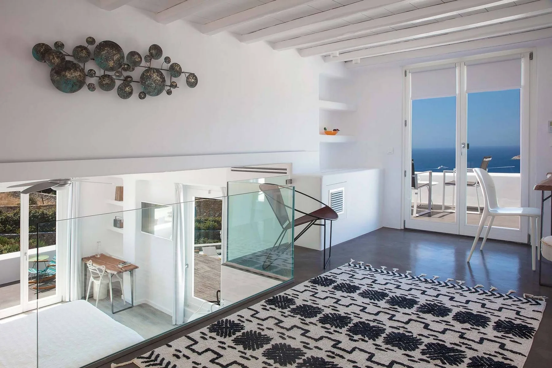 A bright loft sitting area with a patterned rug and glass railing overlooks the sea.