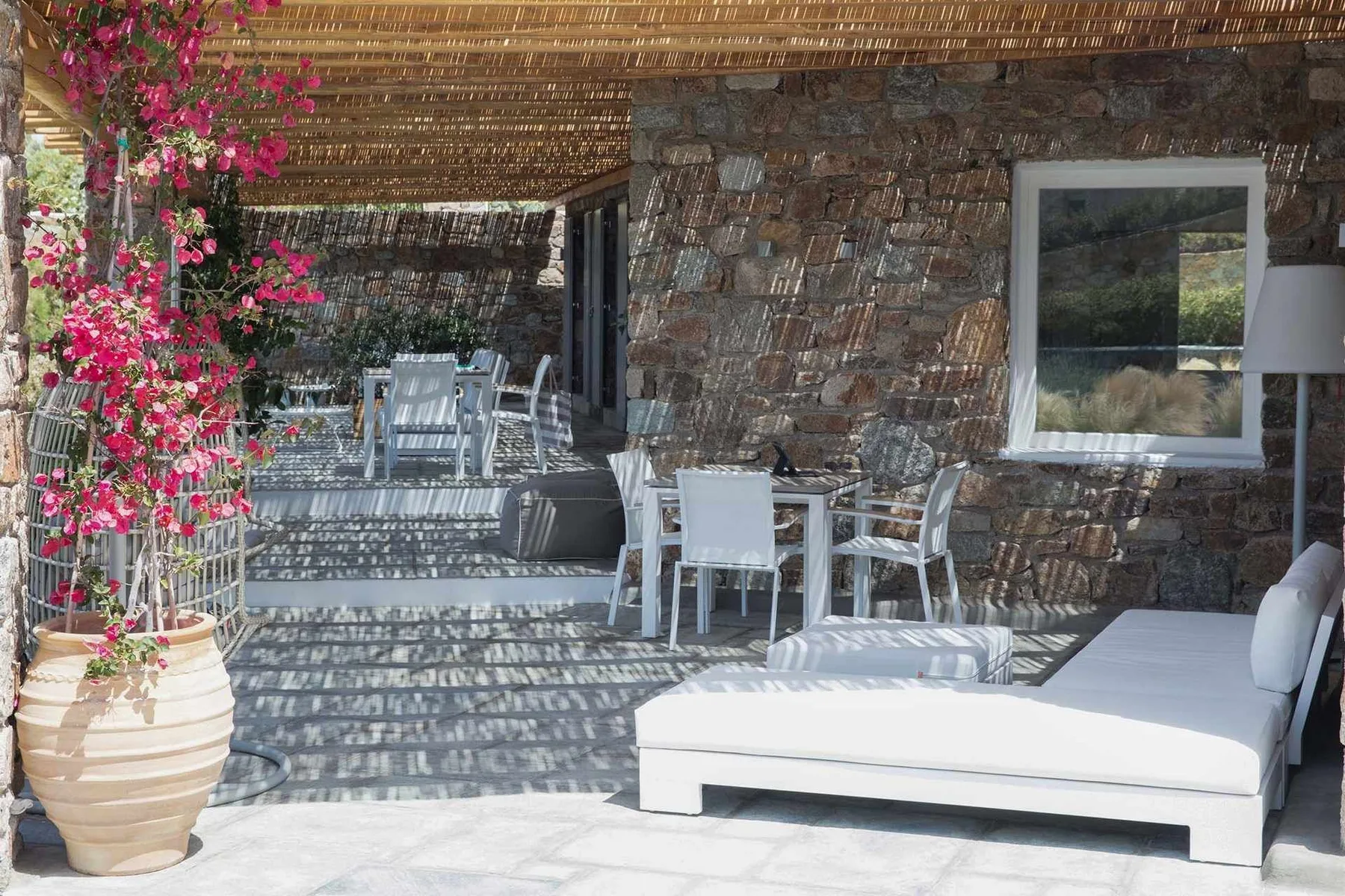 A shaded stone patio has outdoor seating and a white lounge chair.
