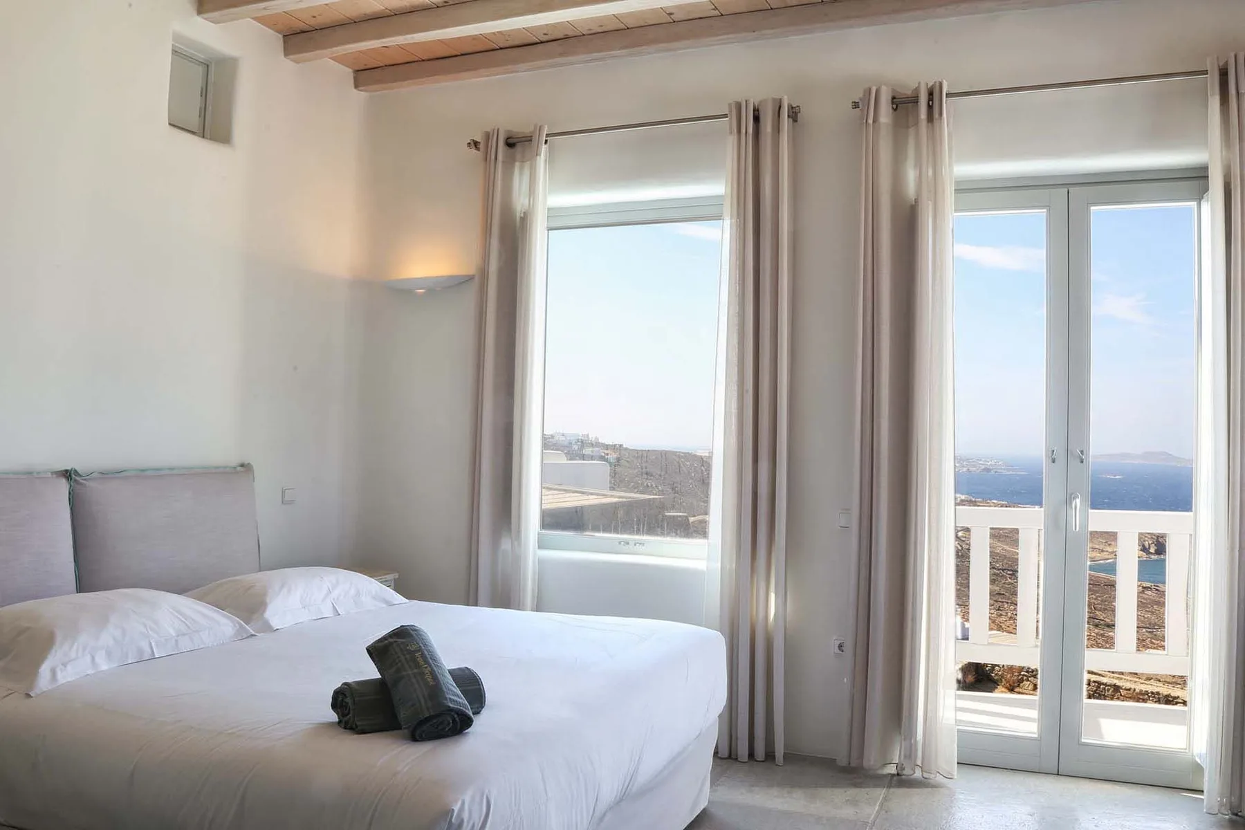 A bright bedroom with a bed beside tall windows overlooking the sea.