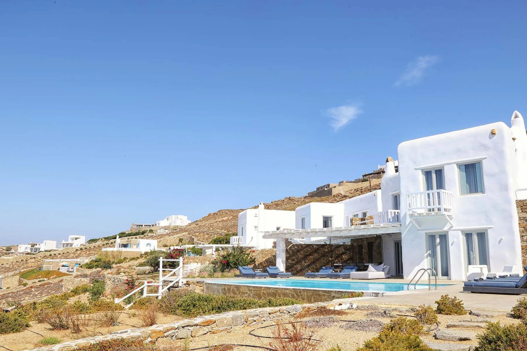 A white modern house sits beside a swimming pool in a dry, open landscape.
