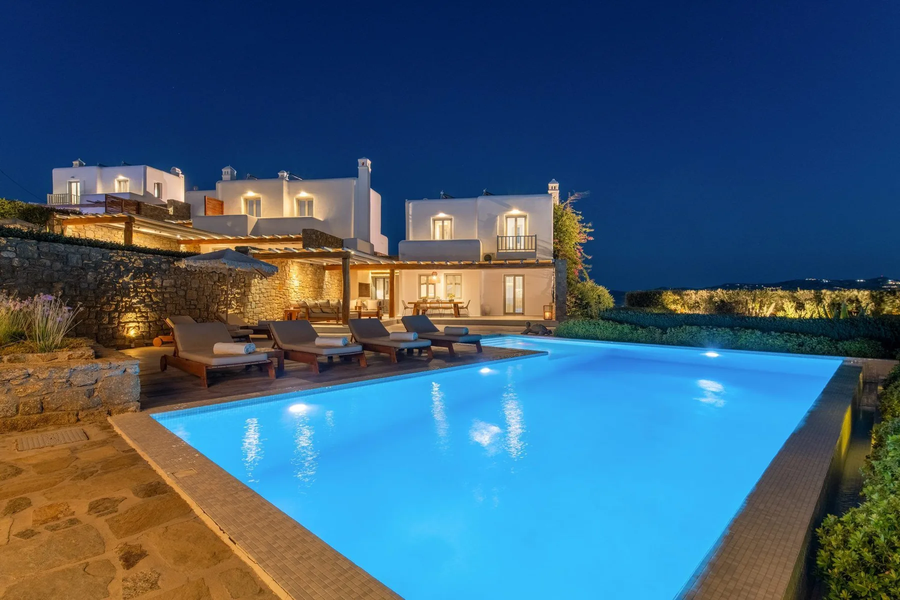A lit outdoor swimming pool sits beside a modern house at night.