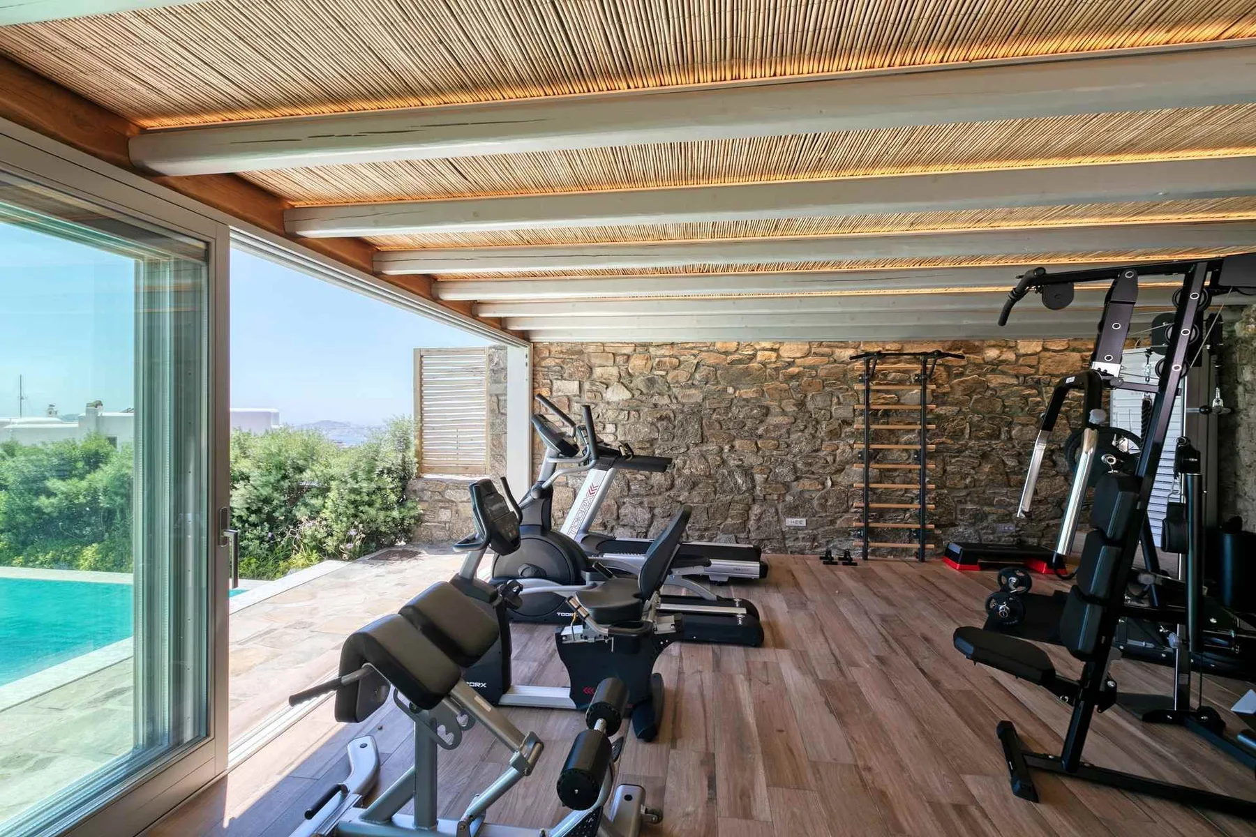 A gym with exercise machines and a stone wall, opening onto a poolside view.