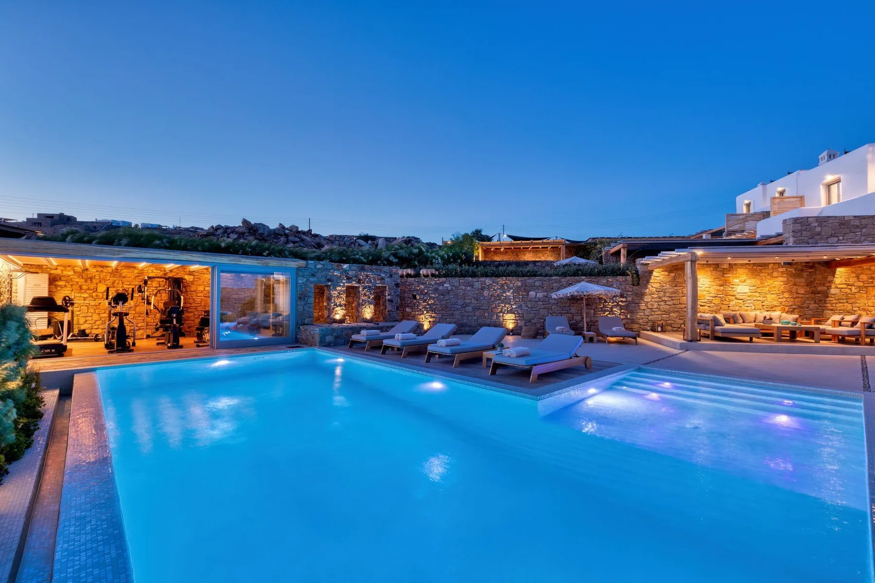 Poolside scene at dusk with illuminated water and seating beside a stone wall.
