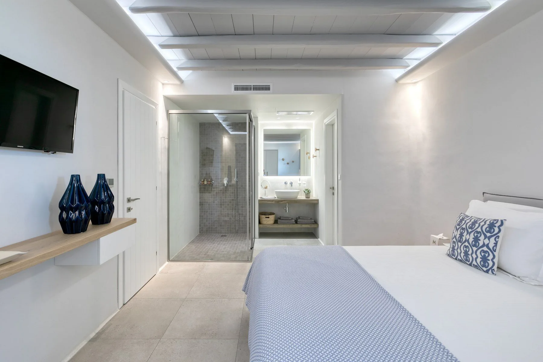 A bright bedroom with a bed beside an open bathroom vanity and shower.